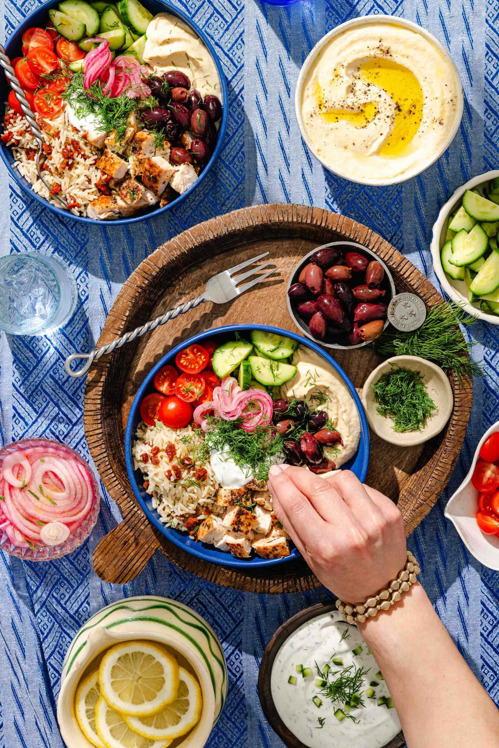 A hand reaches for a bowl of Mediterranean food with rice, grilled chicken, tomatoes, cucumbers, pickled onions, olives, and hummus. Surrounding dishes hold hummus, tzatziki, olives, cucumbers, tomatoes, lemon, and herbs.