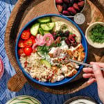 A bowl with grilled chicken, rice, cherry tomatoes, cucumber, olives, pickled onions, and hummus, garnished with dill. A hand holds a fork above the bowl. Side dishes of olives and herbs are nearby on a wooden tray.
