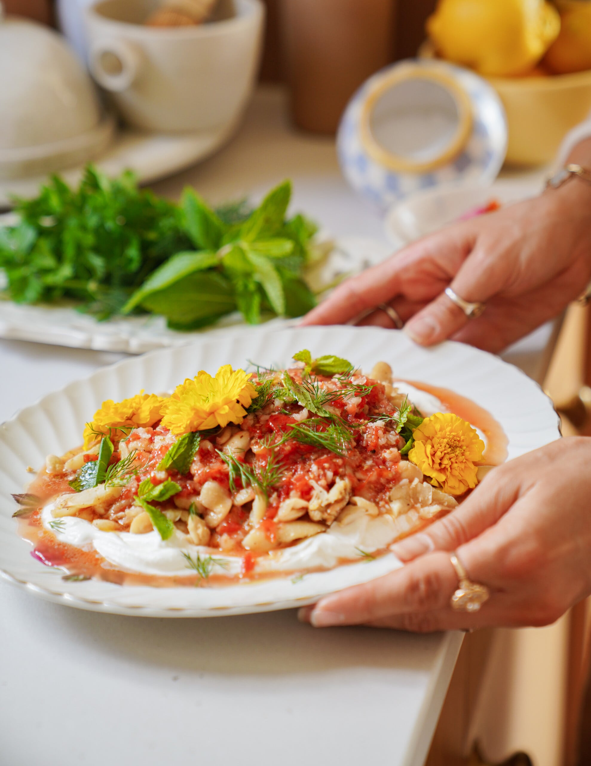 A person holds a plate of food garnished with edible yellow flowers, fresh herbs, and a tomato-based sauce, with bowls of herbs and lemons visible on the counter in the background.