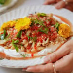 A plate of food with beans, tomato sauce, fresh herbs, and edible yellow flowers on a layer of white cream, held by hands with rings on the fingers.