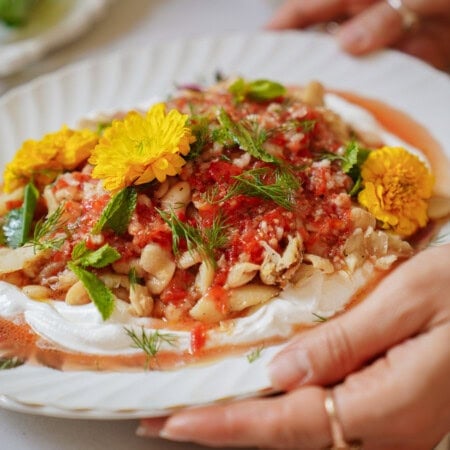 A plate of food with beans, tomato sauce, fresh herbs, and edible yellow flowers on a layer of white cream, held by hands with rings on the fingers.