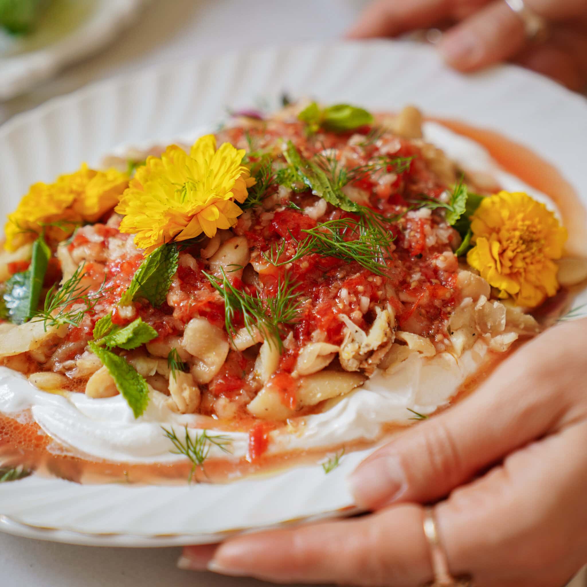 A plate of food with beans, tomato sauce, fresh herbs, and edible yellow flowers on a layer of white cream, held by hands with rings on the fingers.