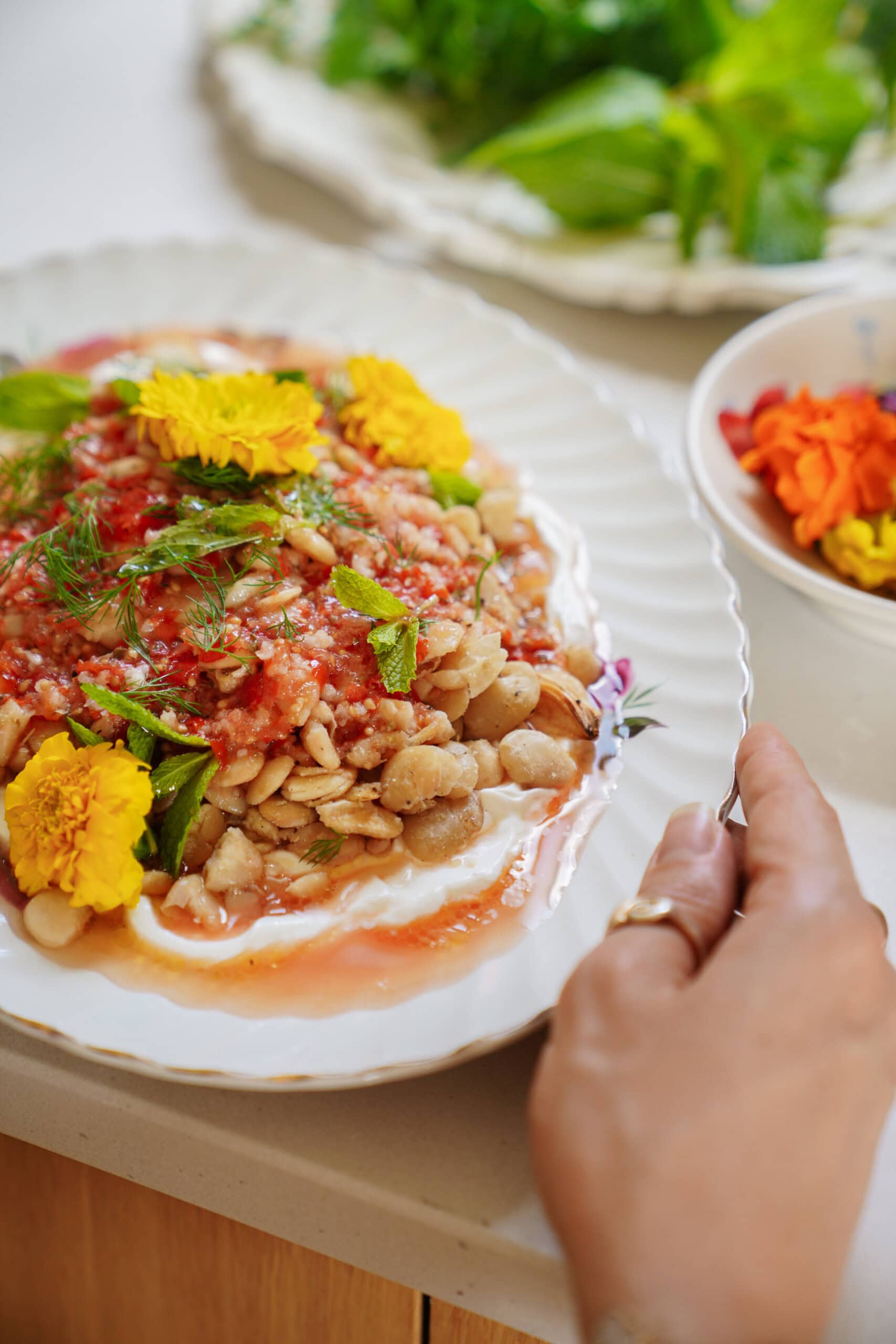 A hand holds a plate of beans topped with tomato sauce, herbs, and edible yellow flowers, with a bowl of marigold petals and a plate of fresh greens in the background.