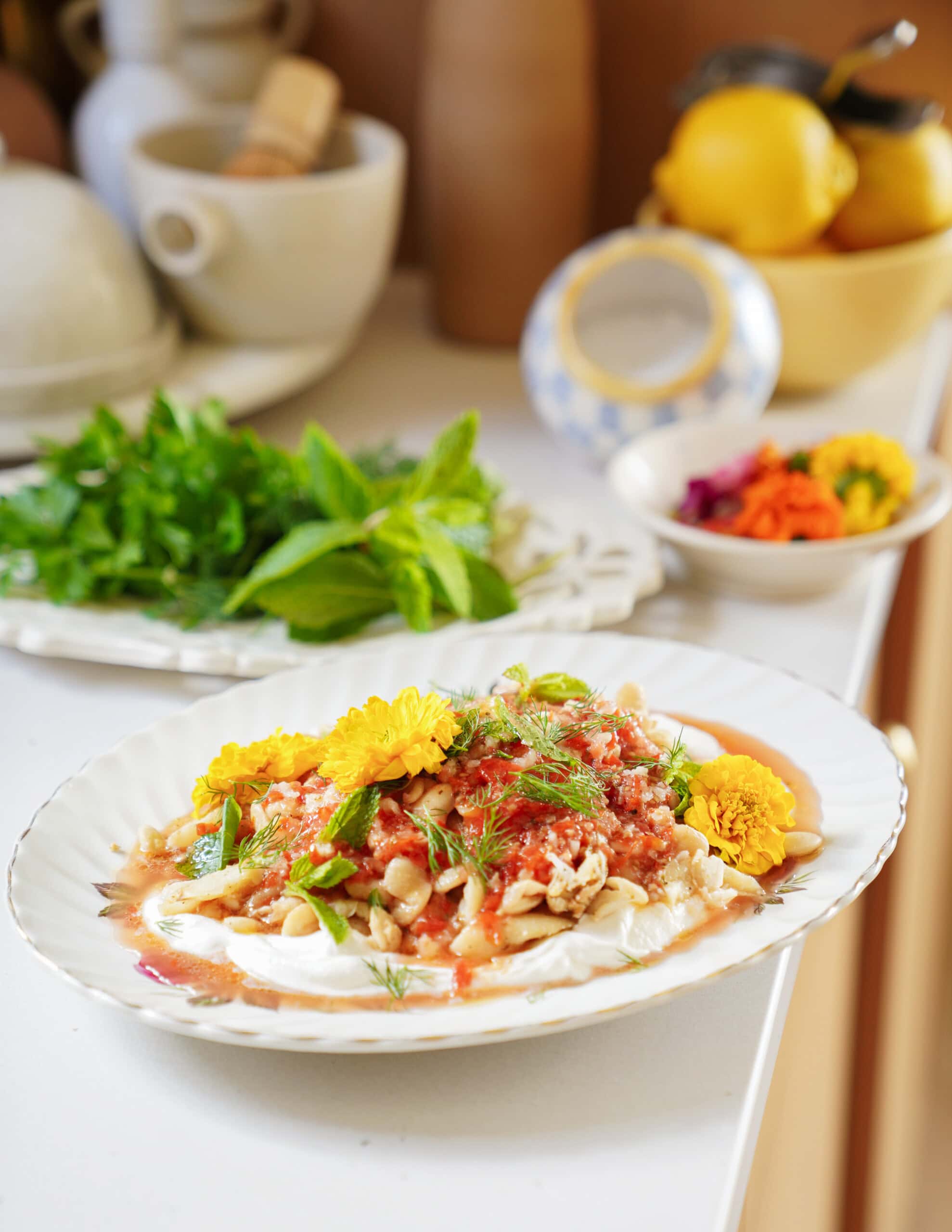 A plate of salad topped with fresh herbs, nuts, sauce, and yellow edible flowers sits on a white countertop. In the background are a bowl of greens, a mortar and pestle, lemons, and colorful ingredients.