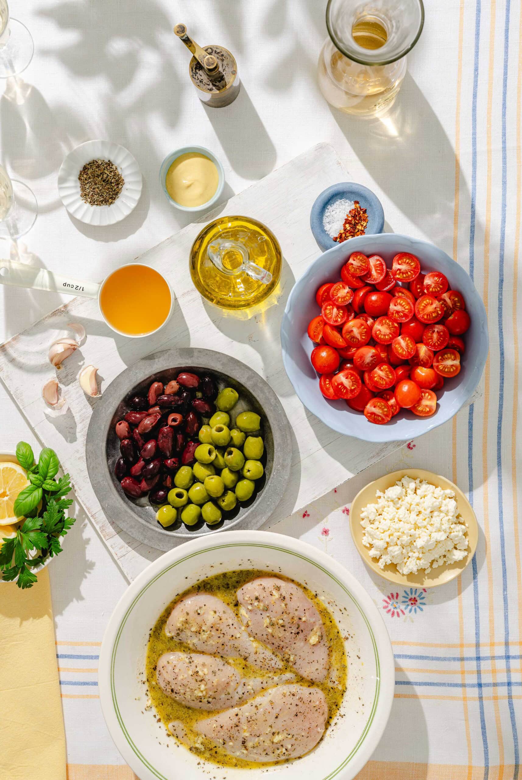 Overhead view of a table with marinated chicken breasts, cherry tomatoes, green and black olives, crumbled feta cheese, olive oil, garlic, herbs, mustard, pepper, orange liquid, and kitchen utensils.