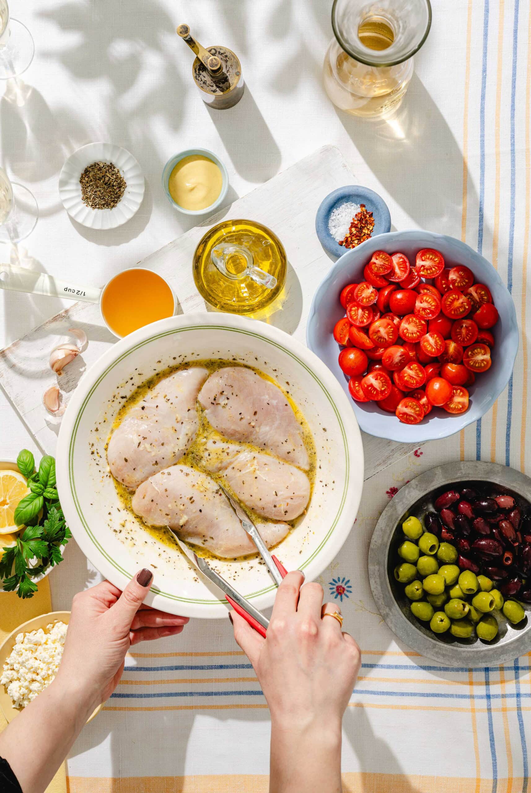 A person uses tongs to marinate raw chicken breasts in a bowl surrounded by cherry tomatoes, olives, feta cheese, olive oil, seasonings, and herbs on a sunlit table.