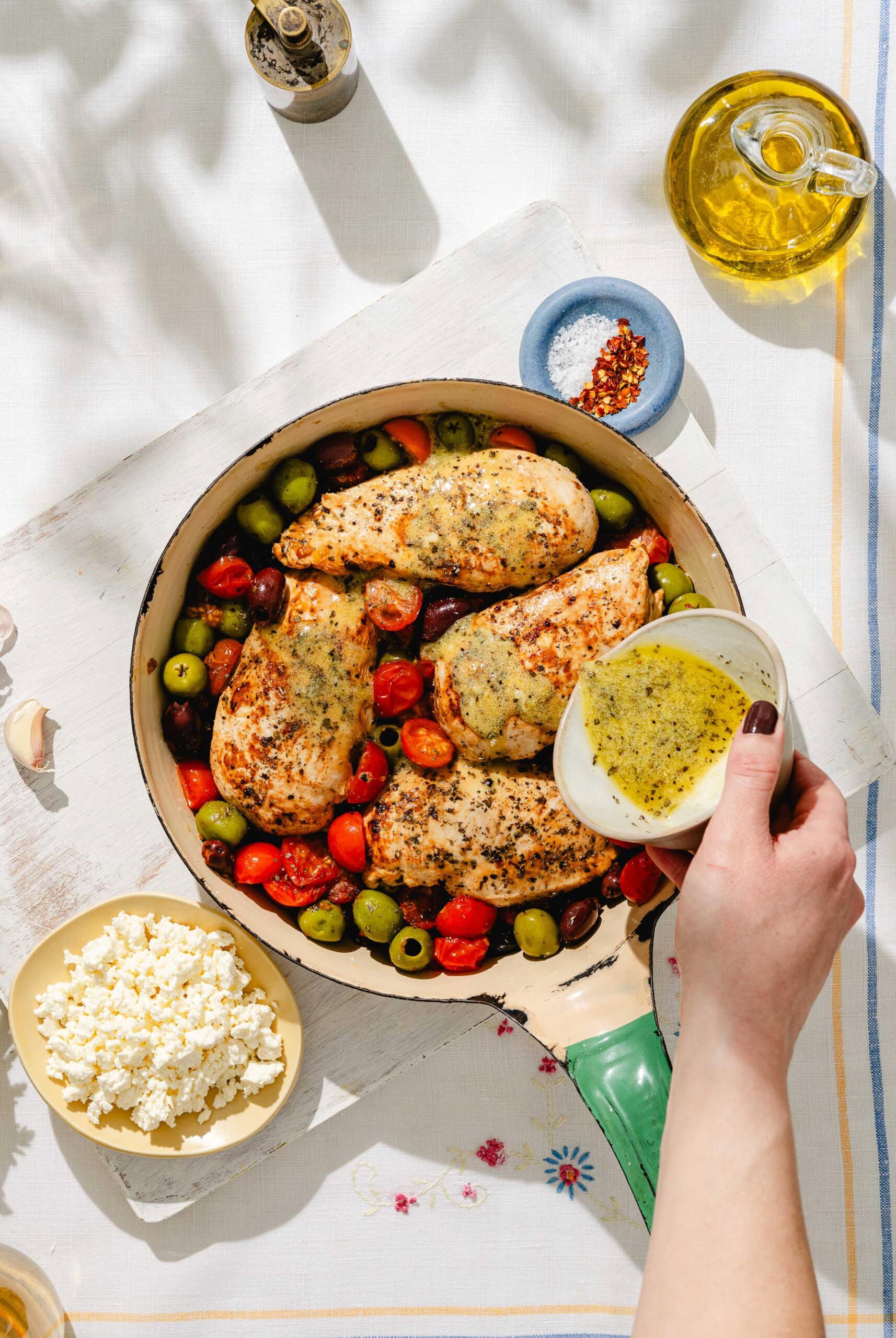 A hand pours a yellow herb sauce over seasoned chicken breasts in a skillet with olives and cherry tomatoes. Nearby are a bowl of crumbled feta, a small bowl of salt, olive oil, and a pepper mill.