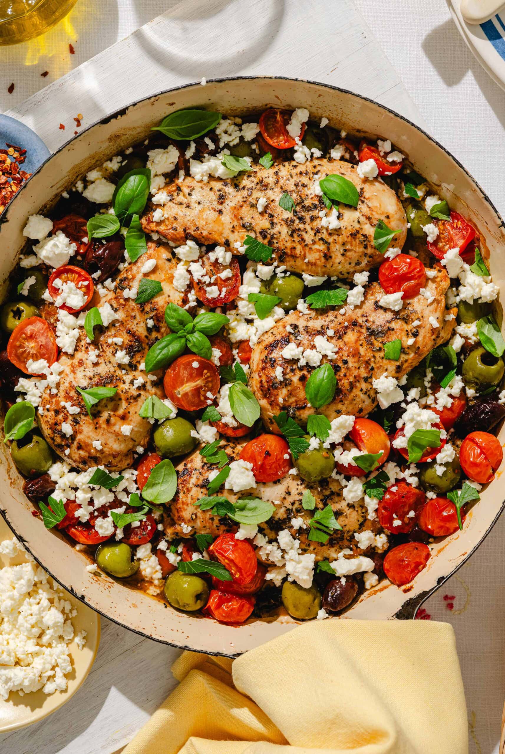 A skillet filled with seasoned chicken breasts, cherry tomatoes, green and black olives, crumbled feta cheese, and fresh basil leaves, all arranged on a white table.