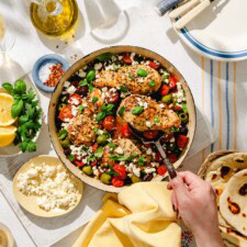 A skillet filled with seasoned chicken breasts, cherry tomatoes, olives, spinach, and crumbled feta cheese is on a table. A hand serves a piece of chicken. Fresh herbs, lemon slices, flatbread, and olive oil are nearby.
