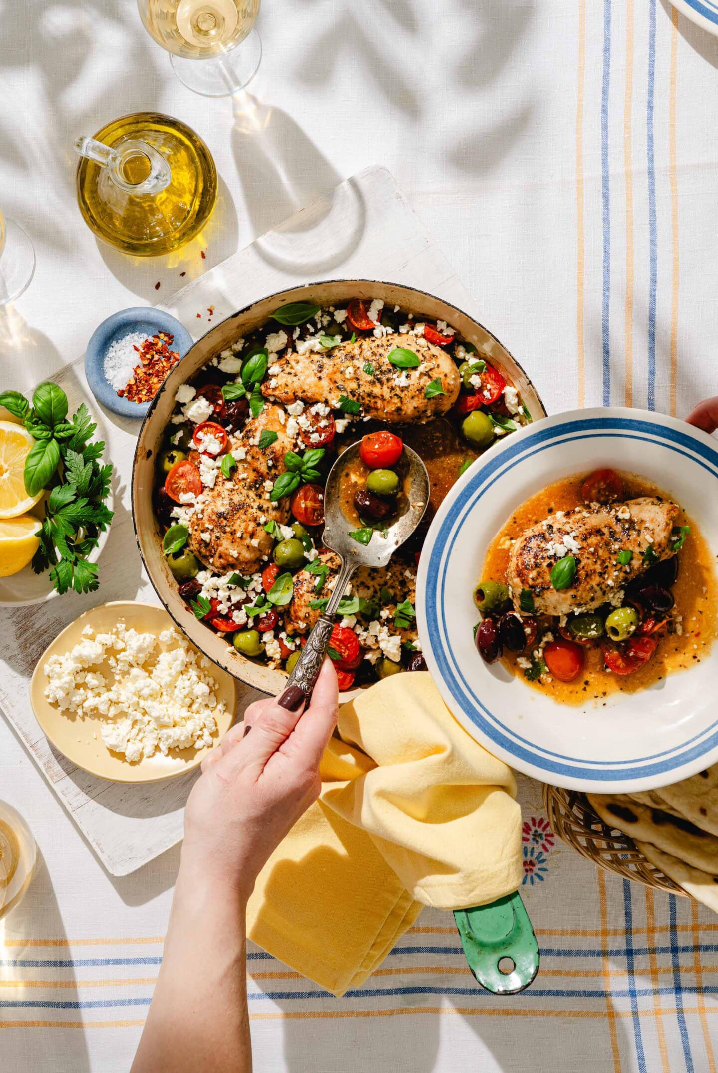 A hand serves Mediterranean chicken with olives, tomatoes, and feta from a skillet into a bowl on a table set with wine, olive oil, lemon, herbs, crumbled feta, and a yellow napkin in bright natural light.