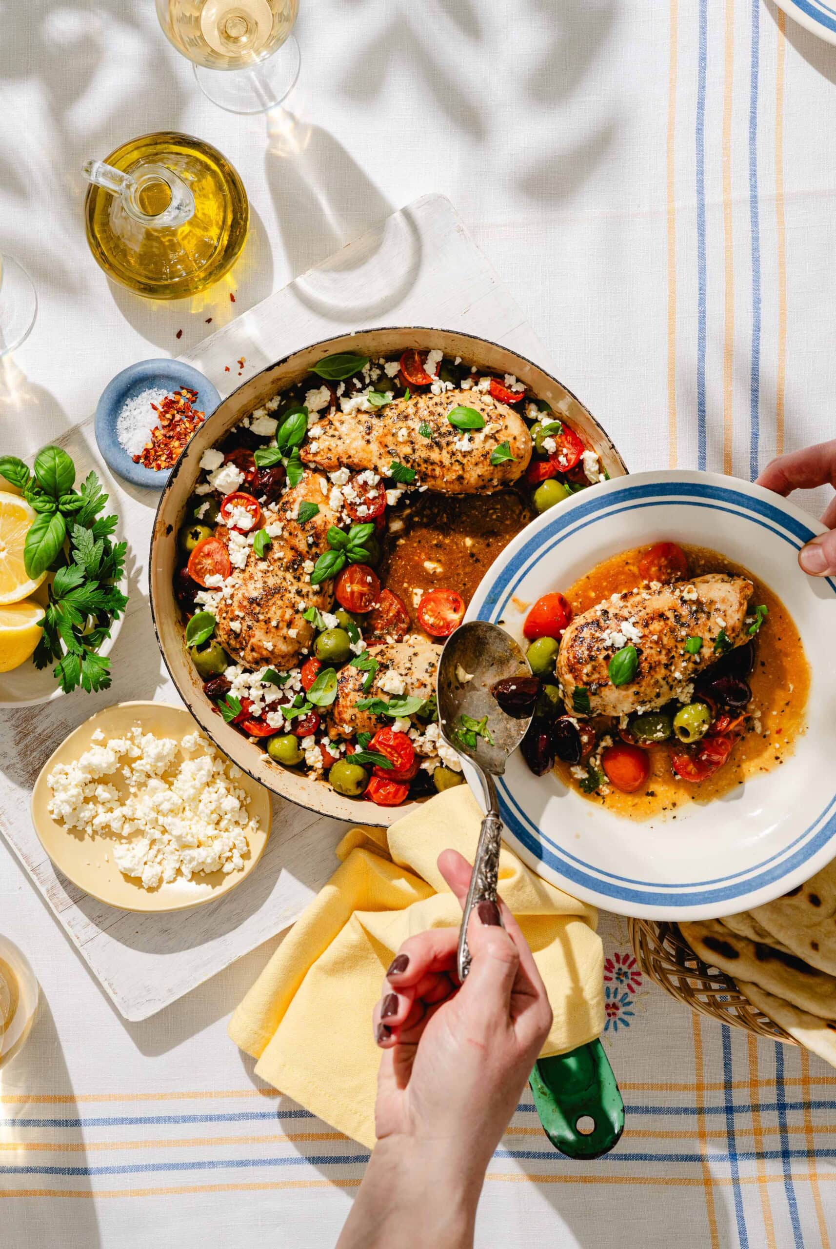 A person serves a portion of baked chicken with tomatoes, olives, feta, and herbs from a skillet onto a plate. The table also has feta cheese, lemon, olive oil, and wine on a striped tablecloth.