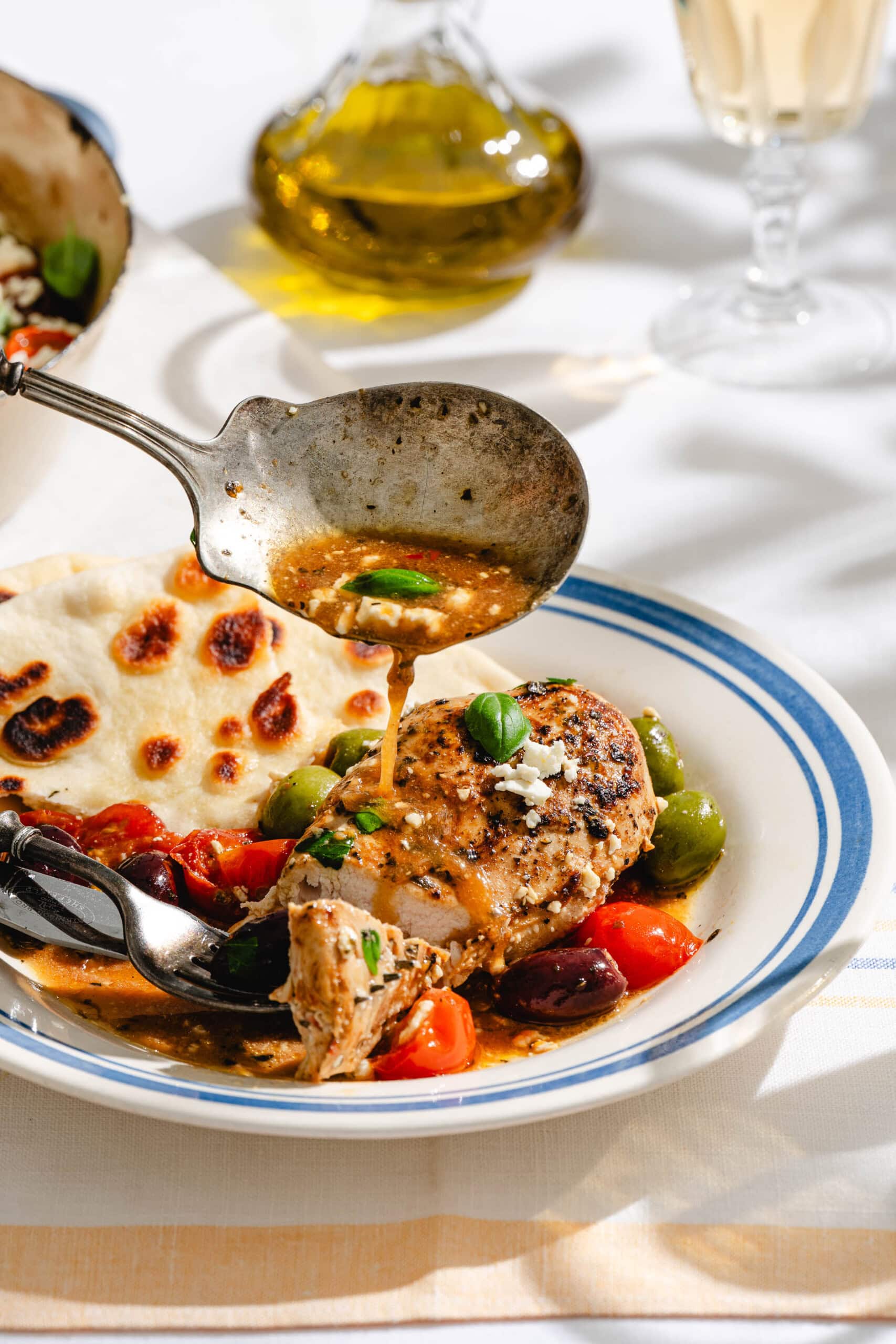 A spoon pours sauce over grilled chicken breast served with olives, cherry tomatoes, flatbread, and fresh basil on a white plate. A glass of white wine and a bottle of olive oil are in the background.