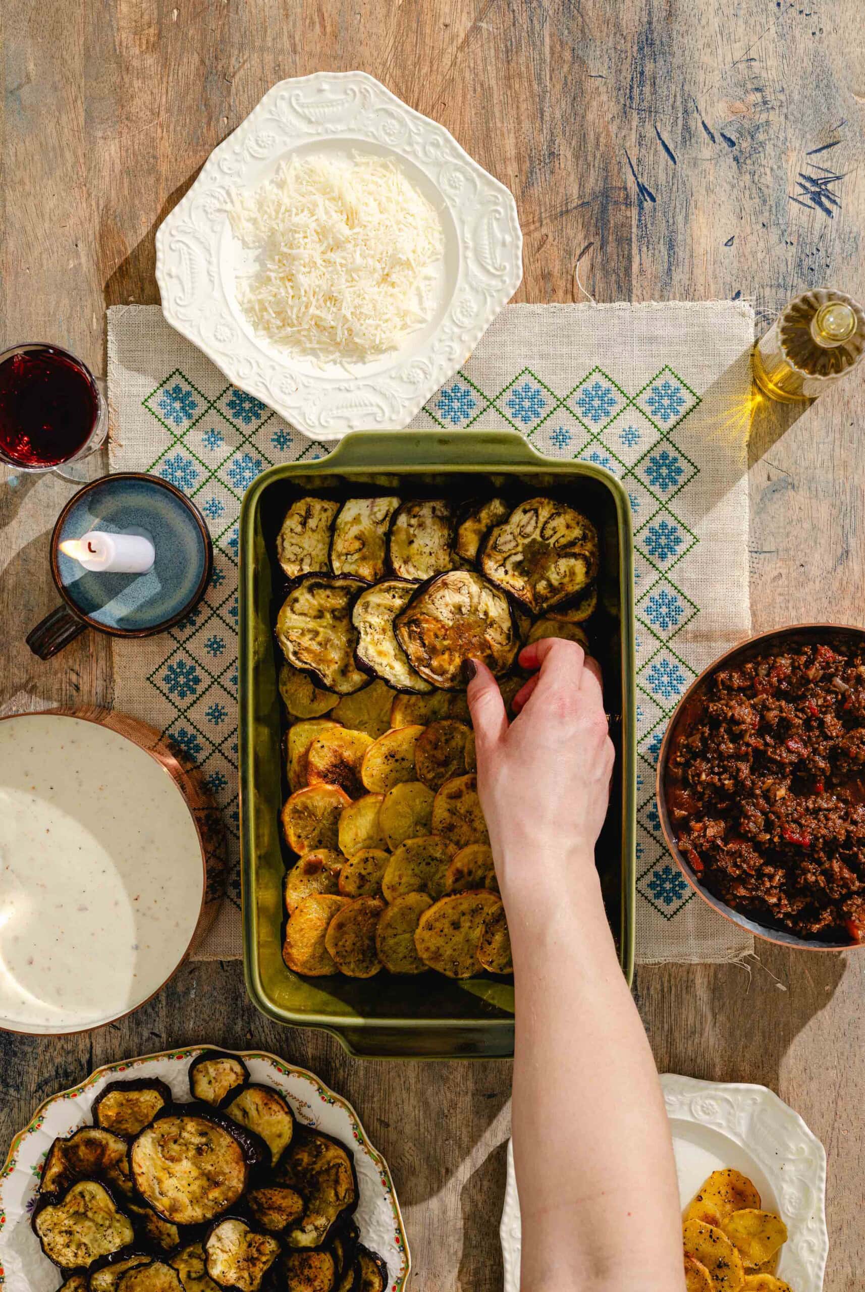 A hand arranges roasted eggplant slices in a baking dish on a wooden table surrounded by plates of cheese, roasted potatoes, meat sauce, a cup, and a bottle of olive oil.