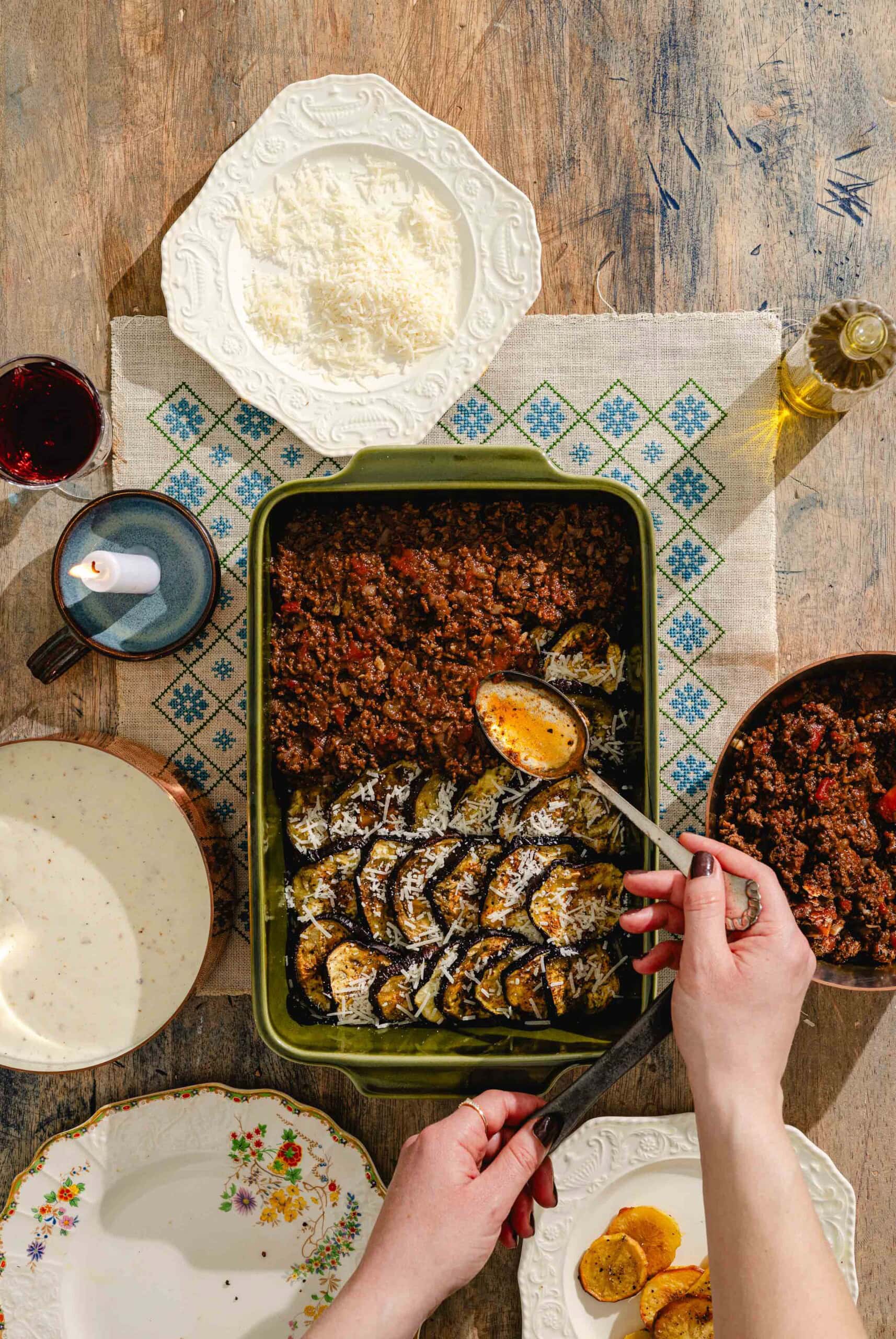 A person assembles a baked dish with sliced eggplant and meat sauce in a green pan on a rustic table, surrounded by plates, grated cheese, a cup, and a bottle of olive oil.