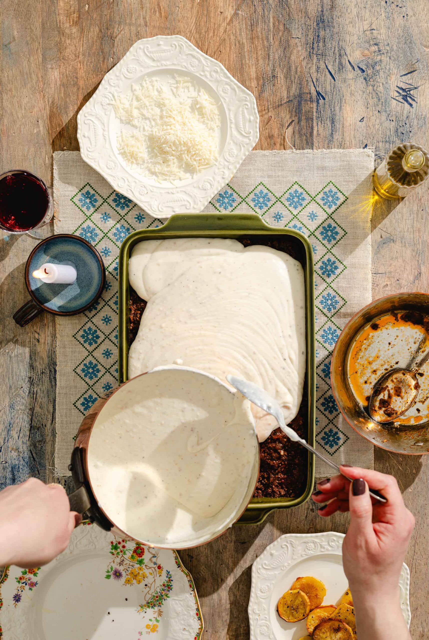 A person pours creamy white sauce from a saucepan over a baking dish with a brown layer, surrounded by plates, a glass of wine, olive oil, grated cheese, and a bowl on a rustic wooden table.
