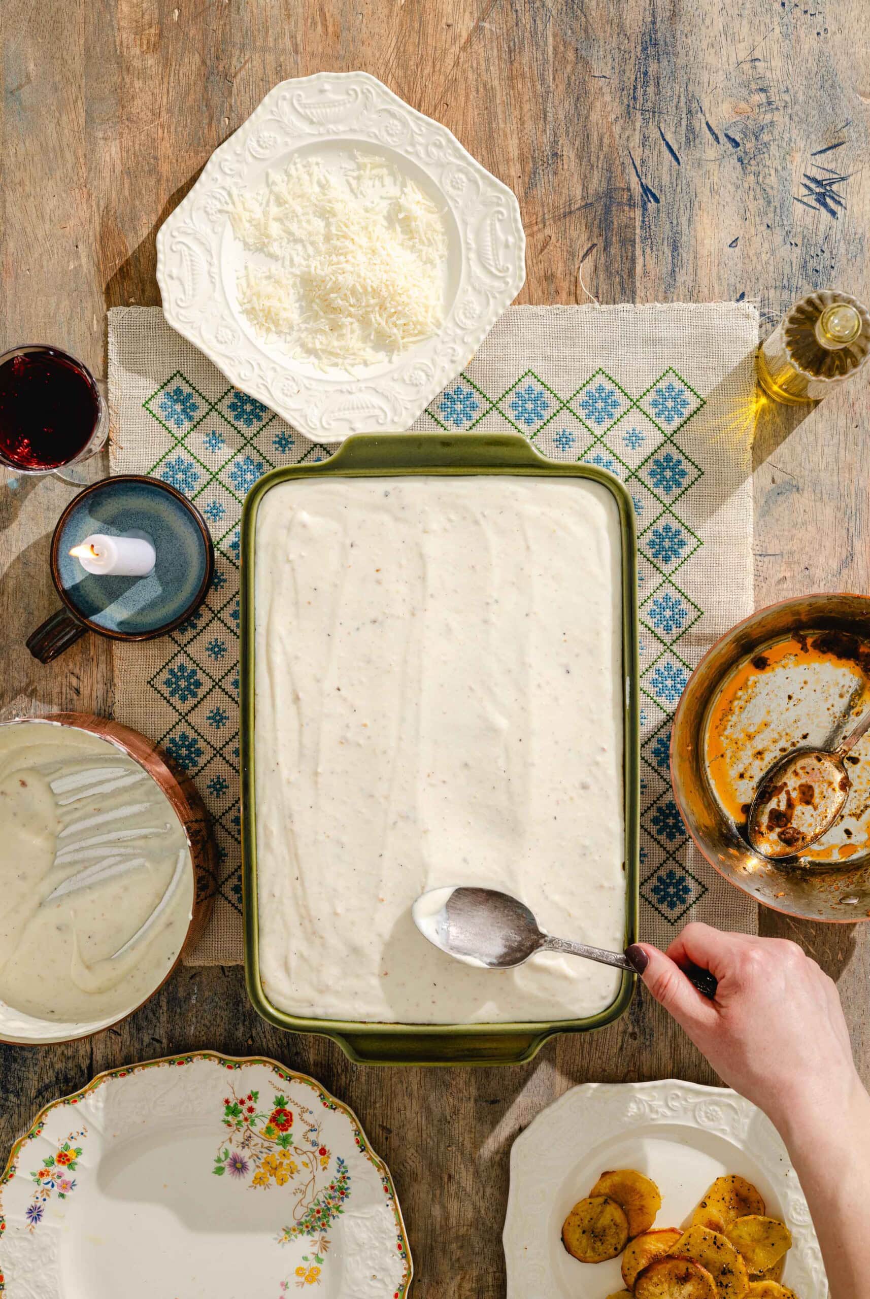A hand spreads creamy white sauce over a dish in a green baking pan. Surrounding the pan are plates with grated cheese, empty dishes, a small cup, a bowl with sauce, slices of cooked potato, and a glass of red wine.