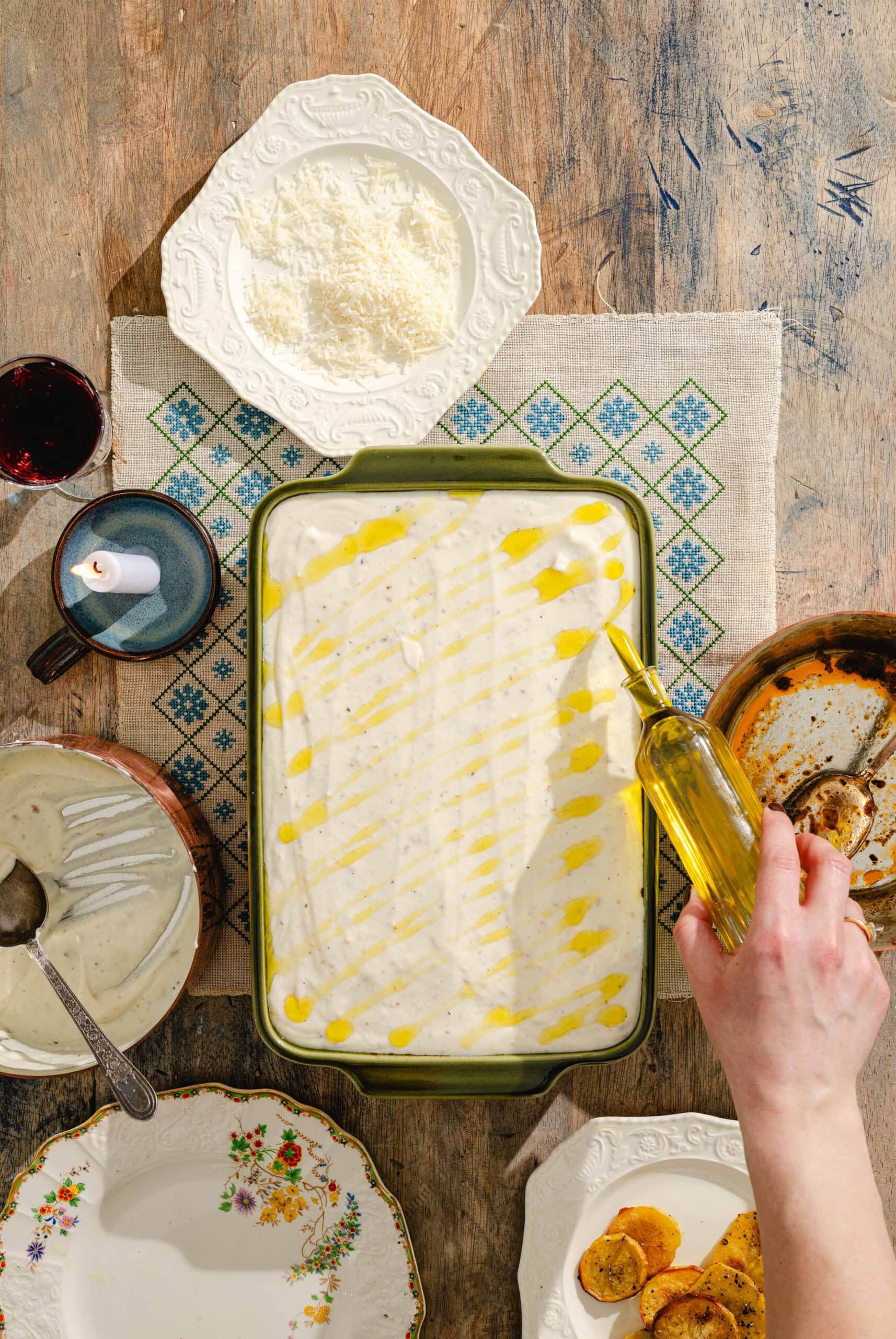 A hand drizzles olive oil over a casserole dish filled with a creamy, white-topped dish on a wooden table surrounded by plates, a bowl of cheese, a cup of coffee, wine, and roasted vegetables.