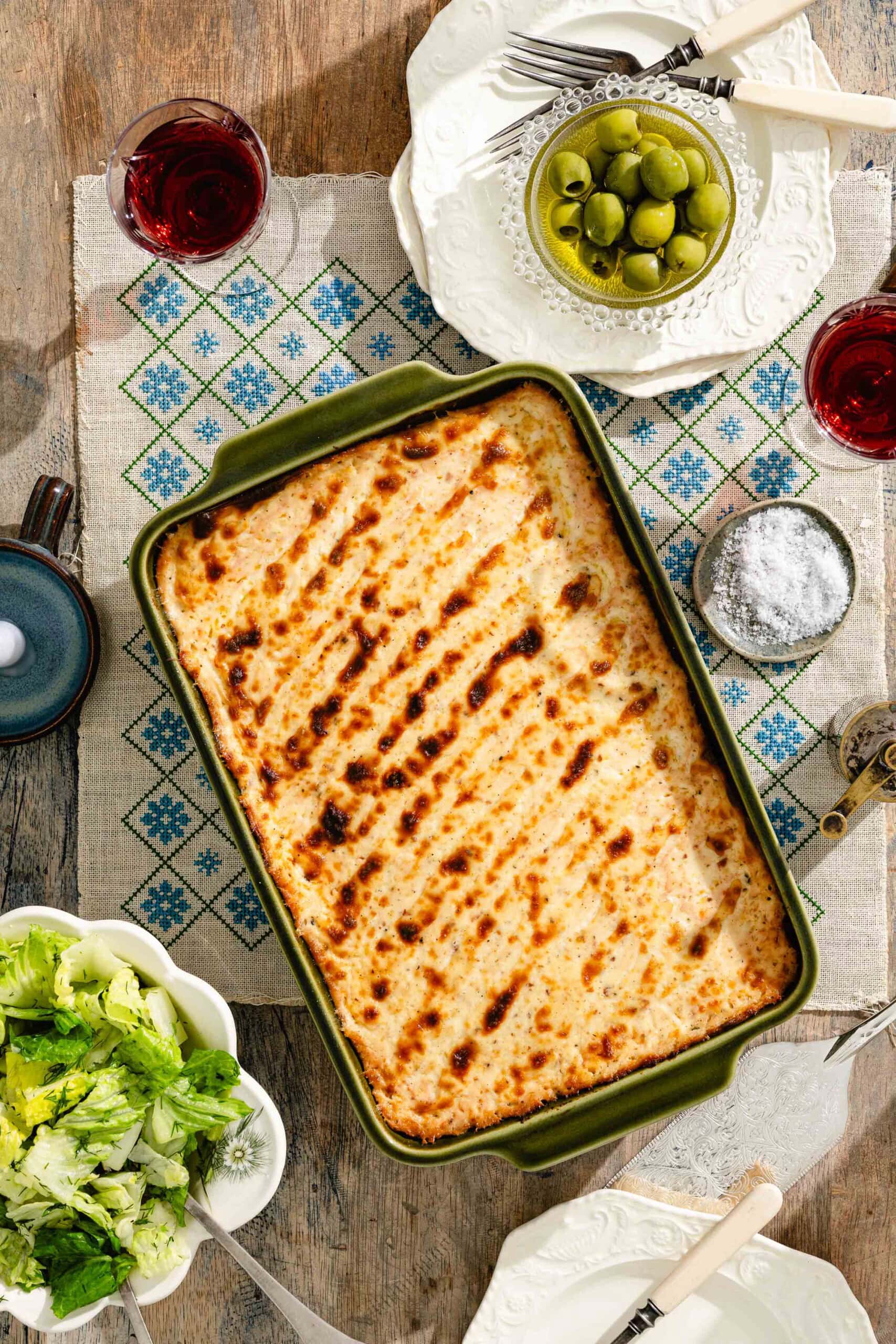 A baked casserole with a golden, browned top sits on a patterned cloth, surrounded by plates with a green salad, a bowl of green olives, a dish of salt, and glasses of red drink on a wooden table.