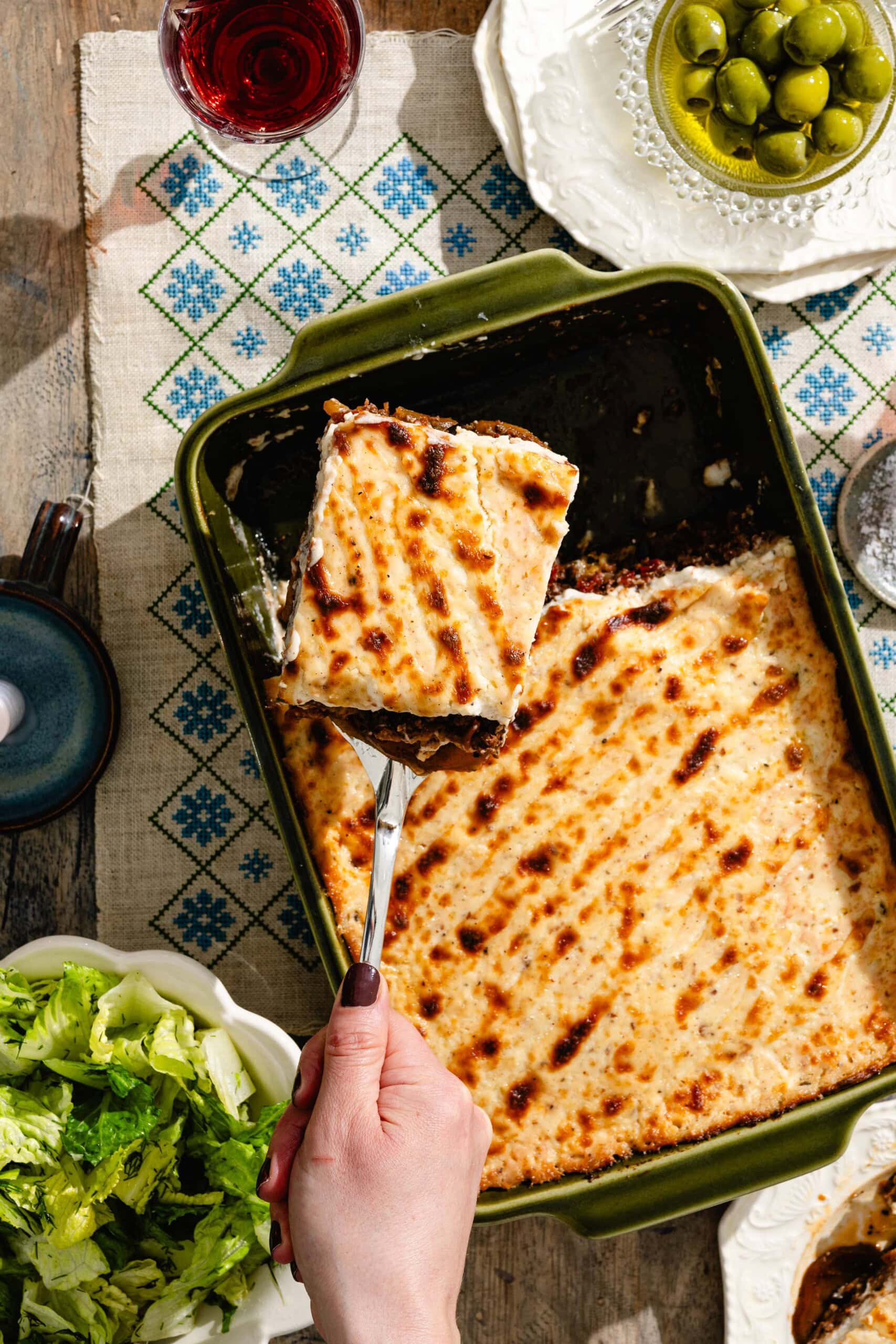 A hand lifts a square serving of baked moussaka from a green dish. The moussaka has a golden-brown topping. Surrounding the dish are a bowl of green olives, a plate of lettuce, and a glass of red wine.
