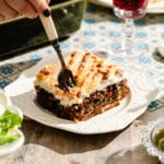 A hand holds a fork over a slice of moussaka on a white plate, with layers of meat, vegetables, and creamy sauce. The dish sits on a rustic table with patterned tablecloth and salad in the background.