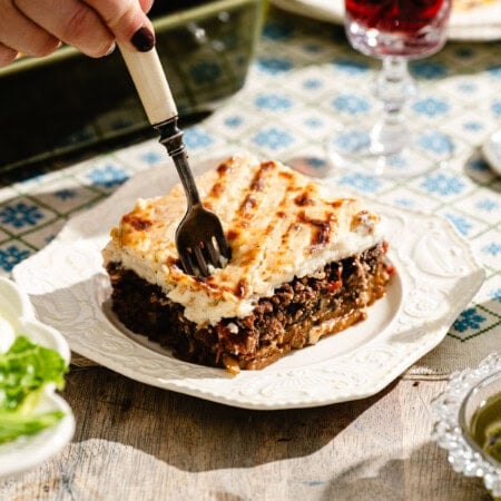 A hand holds a fork over a slice of moussaka on a white plate, with layers of meat, vegetables, and creamy sauce. The dish sits on a rustic table with patterned tablecloth and salad in the background.
