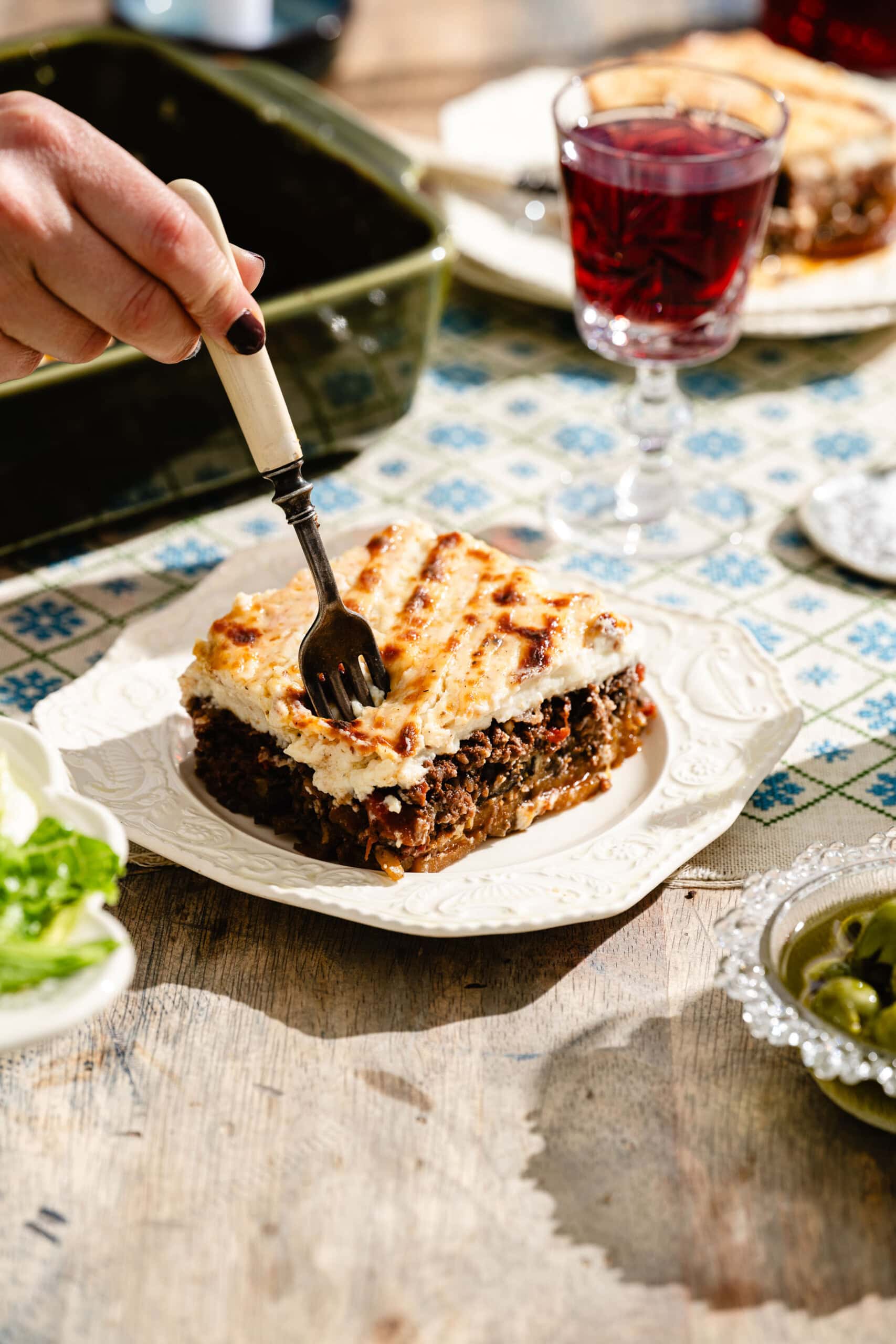 A hand holding a fork approaches a serving of moussaka on a white plate, with a glass of red wine and salad on a patterned tablecloth in bright sunlight.