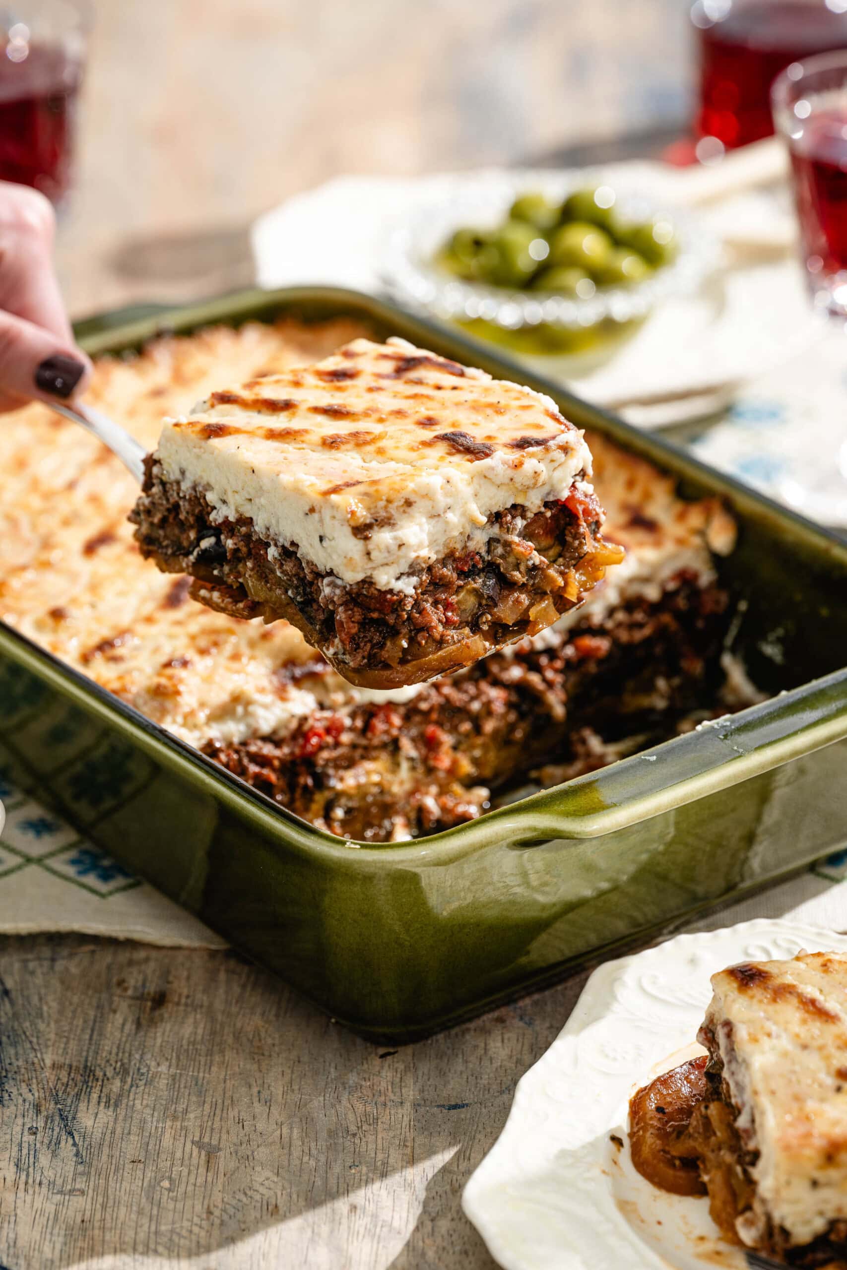 A hand lifts a square serving of baked moussaka from a green casserole dish, revealing layers of meat, vegetables, and creamy topping. A plate with food, green olives, and glasses of red drink are in the background.
