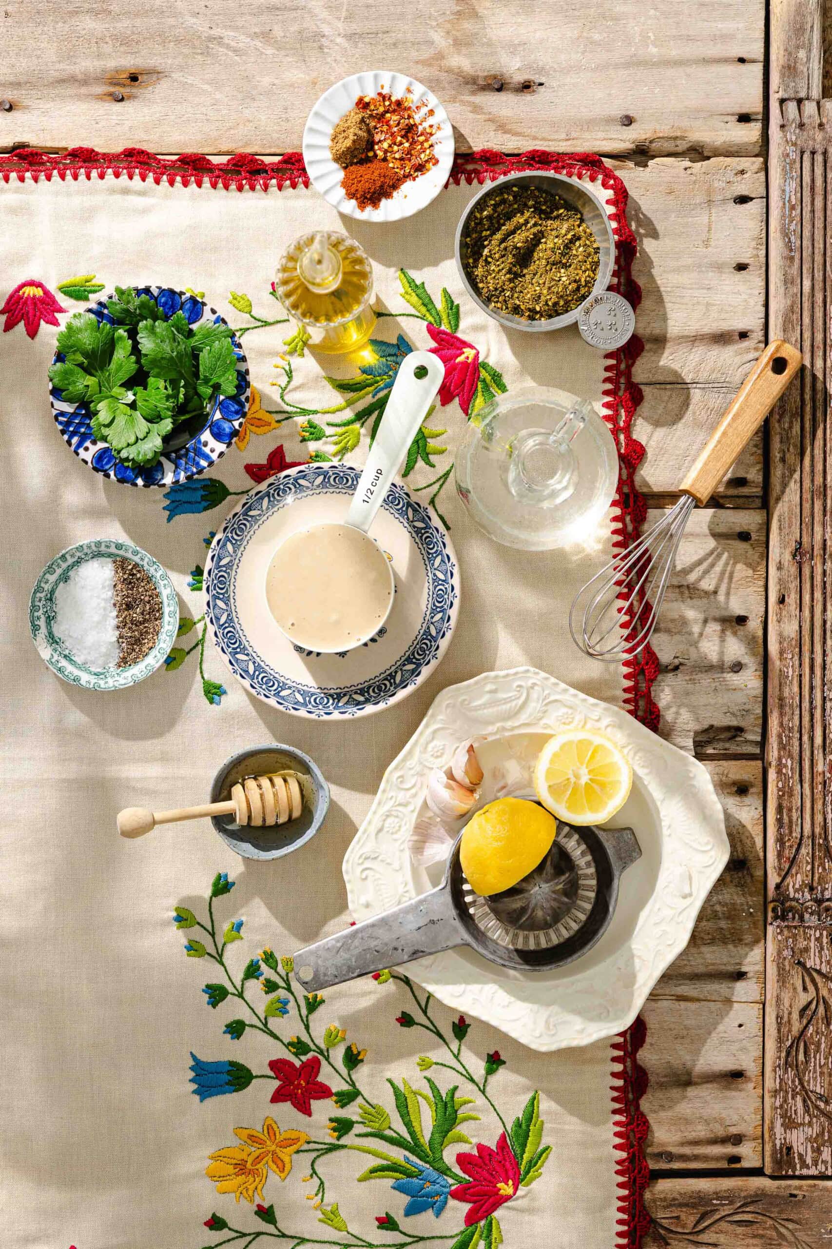 Overhead view of ingredients on a floral tablecloth: fresh herbs, oil, spices, salt, pepper, honey, tahini in a bowl, garlic, lemon halves on a juicer, and a whisk on a rustic wooden table.