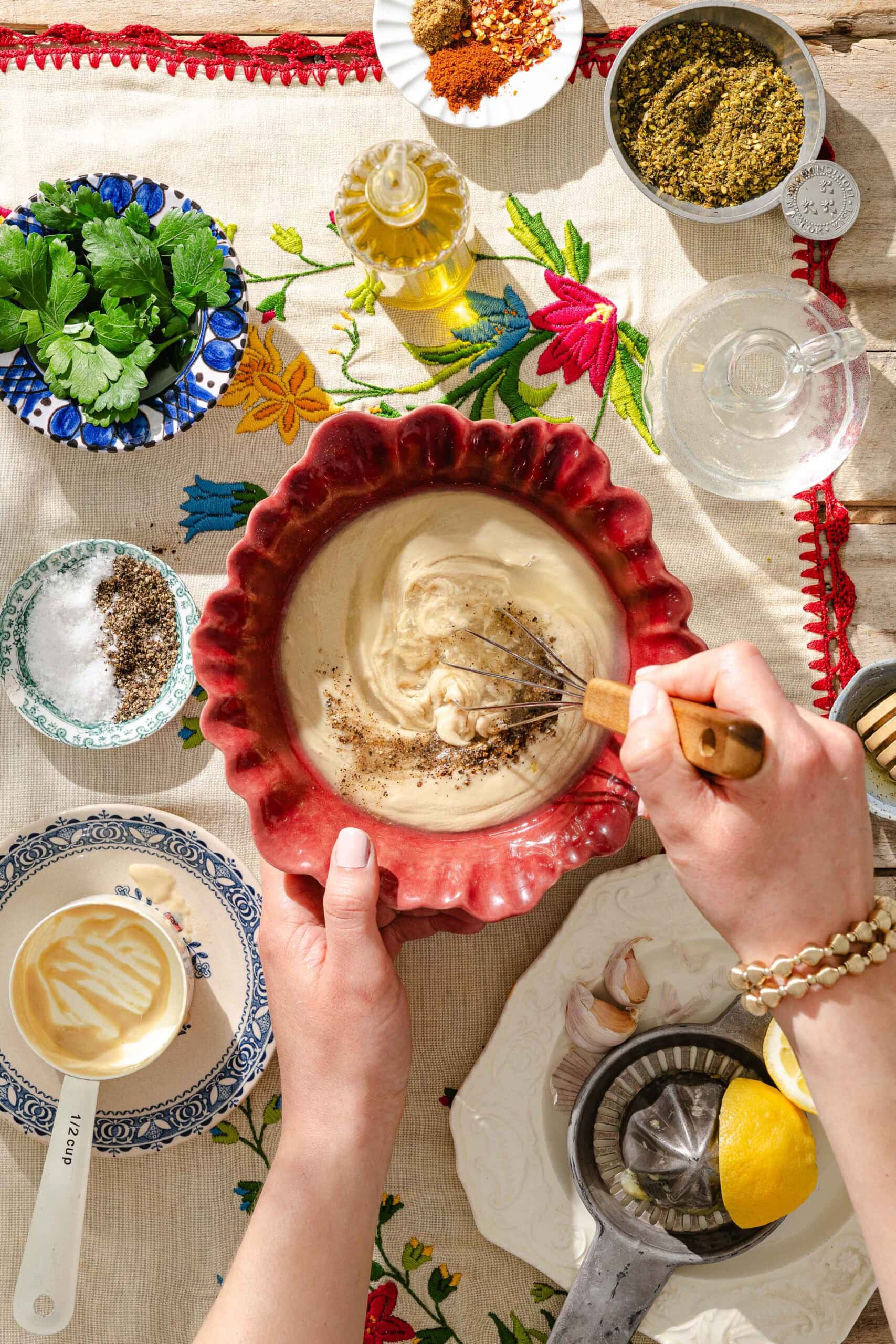 A person whisks tahini sauce in a red bowl on a floral tablecloth, surrounded by fresh herbs, spices, olive oil, lemon, garlic, and small bowls of salt, pepper, and seasonings.