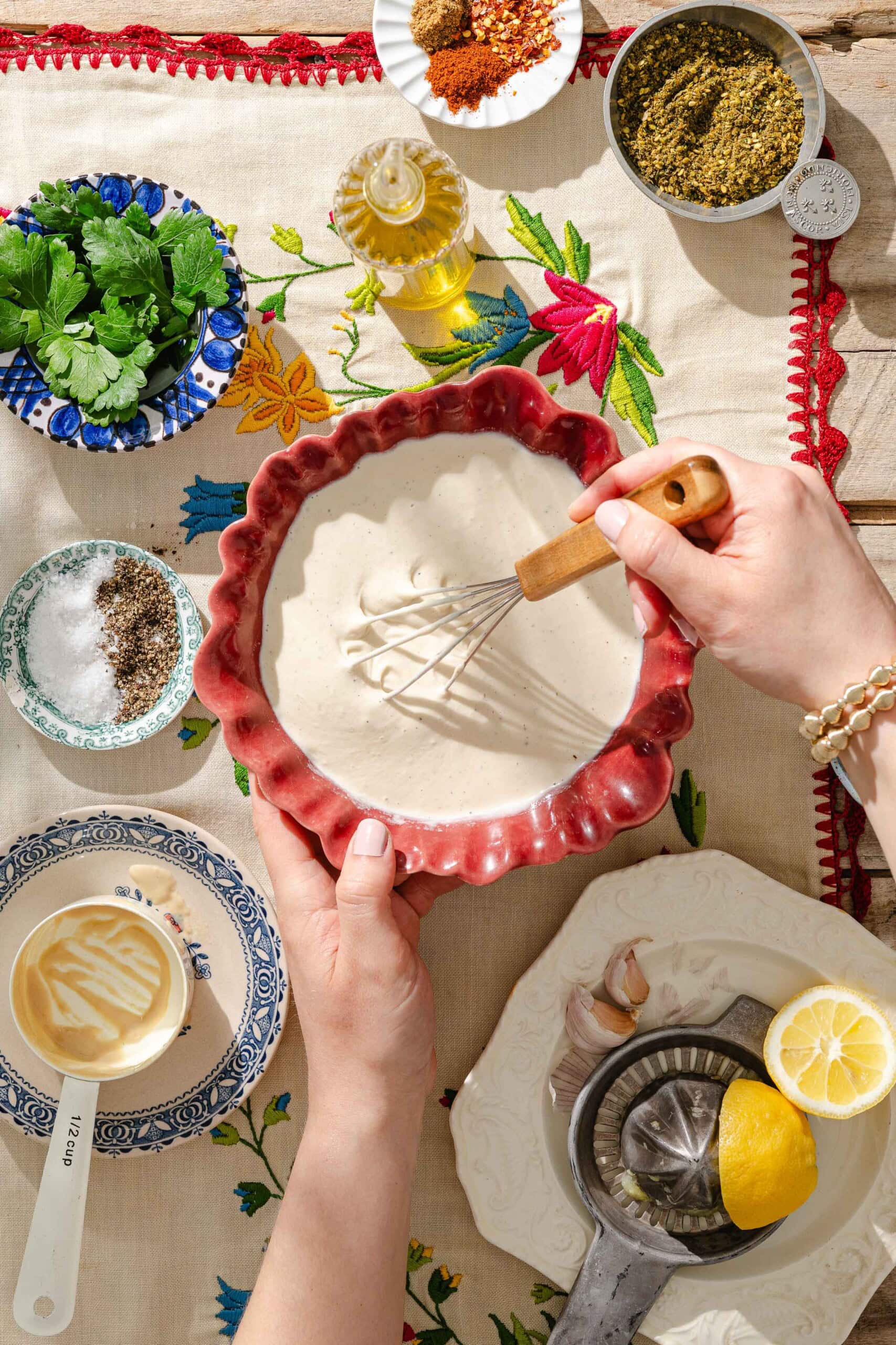 A person whisking tahini sauce in a red bowl surrounded by fresh herbs, spices, olive oil, lemon, garlic, salt, pepper, and a measuring cup on a floral tablecloth.