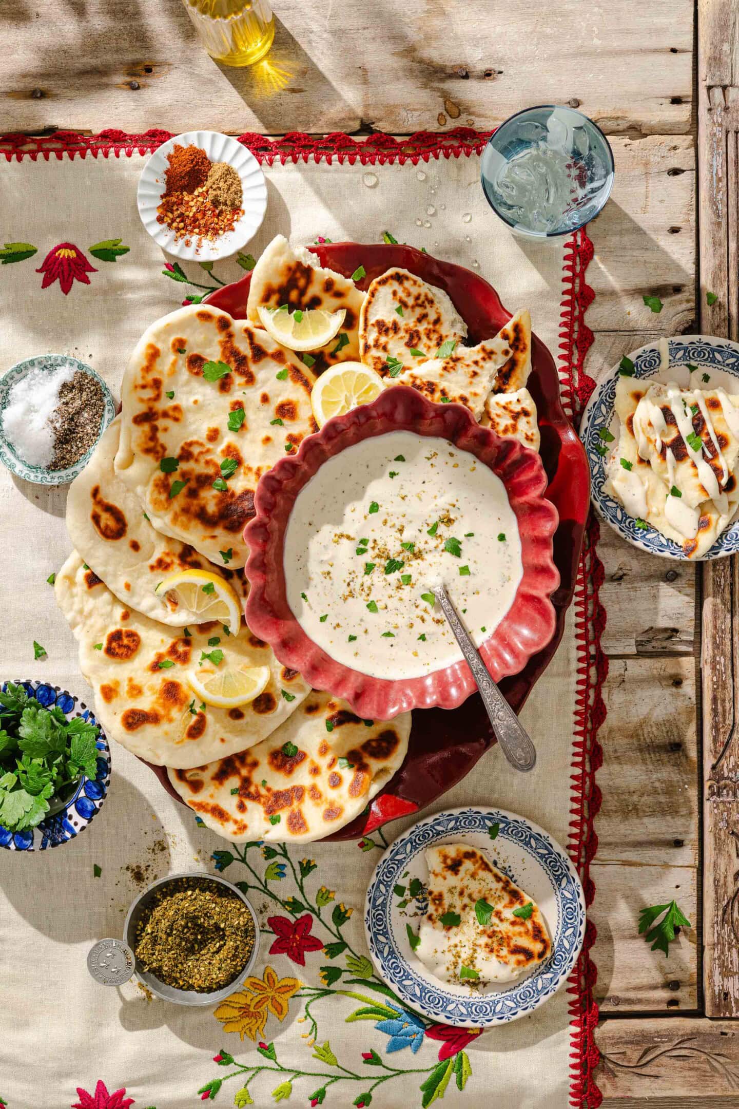 A vibrant spread with naan bread, lemon slices, a large bowl of creamy dip garnished with herbs, and several small bowls containing spices, herbs, and sauce on a floral tablecloth and rustic wooden table.