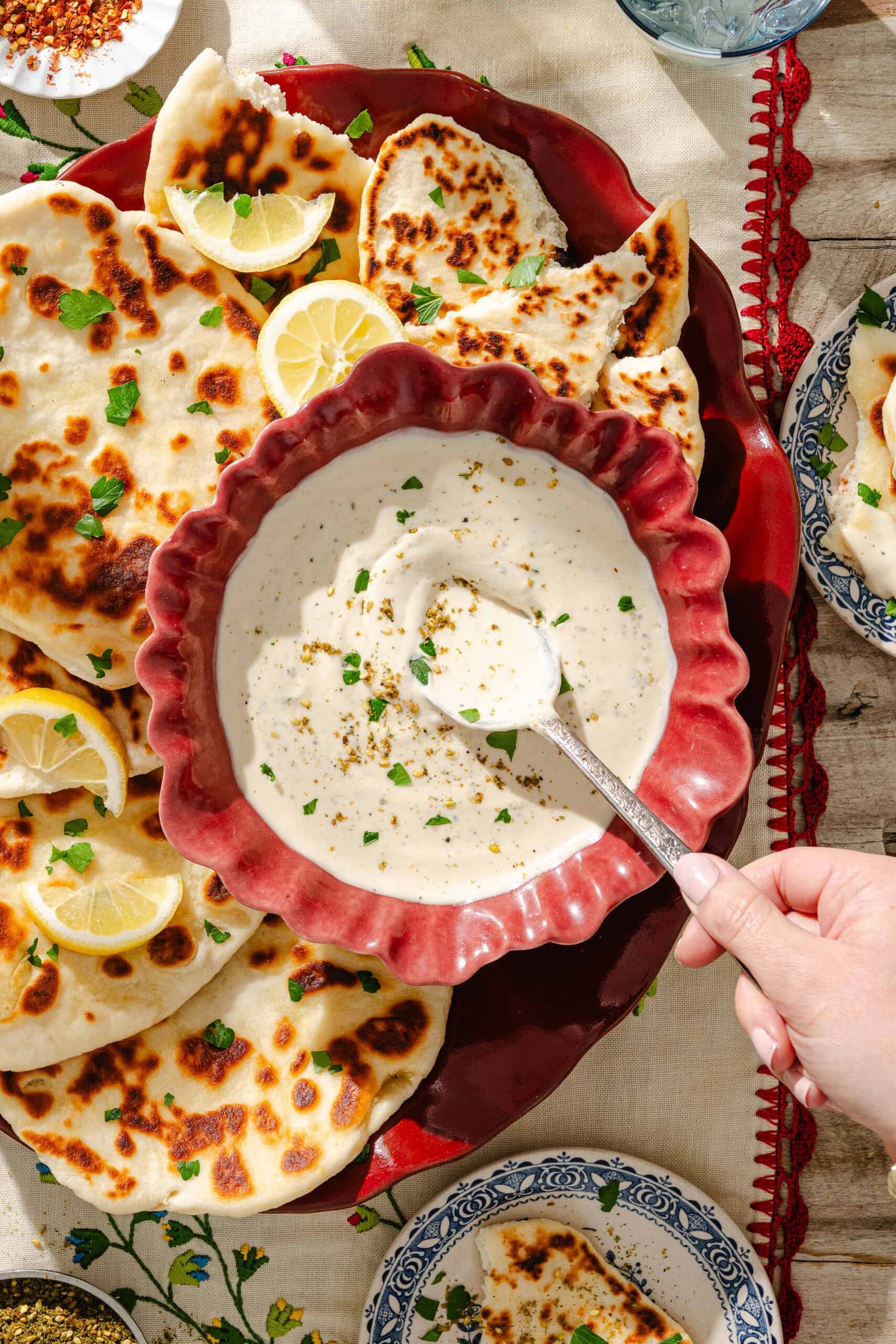 A hand holding a spoon dips into a bowl of creamy white sauce, surrounded by pieces of naan bread, lemon wedges, and herbs on a decorative tablecloth.