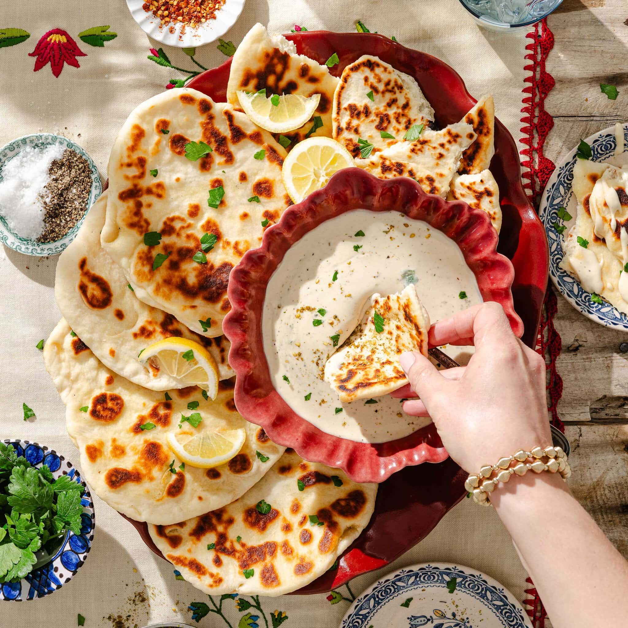 A hand dips a piece of naan bread into a bowl of creamy sauce, surrounded by more naan, lemon slices, herbs, and small dishes of spices on a decorative tablecloth.
