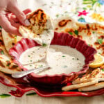 A hand dips a piece of naan bread into a bowl of creamy white sauce, with more naan and a spoon nearby on a floral-patterned cloth.