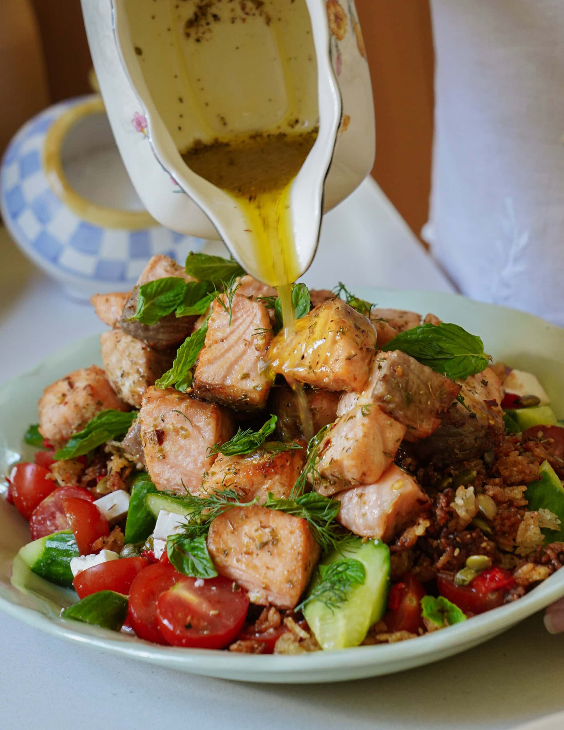 A person pours dressing from a floral-patterned jug onto a fresh salad with grilled salmon, cherry tomatoes, cucumbers, herbs, feta cheese, and greens on a plate.
