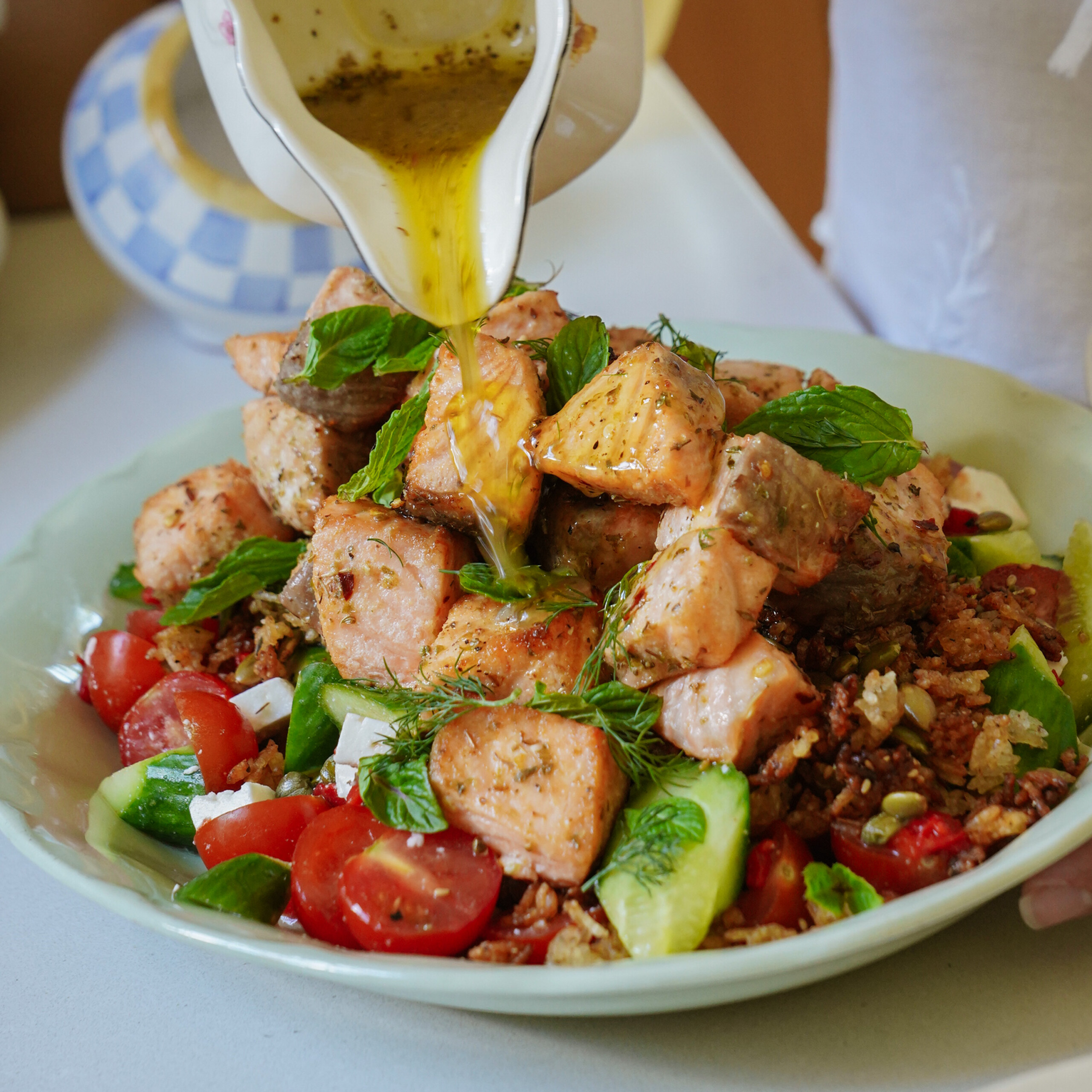 A hand pours olive oil dressing over a salad topped with grilled salmon, cherry tomatoes, cucumber slices, greens, and crumbled cheese on a light green plate.