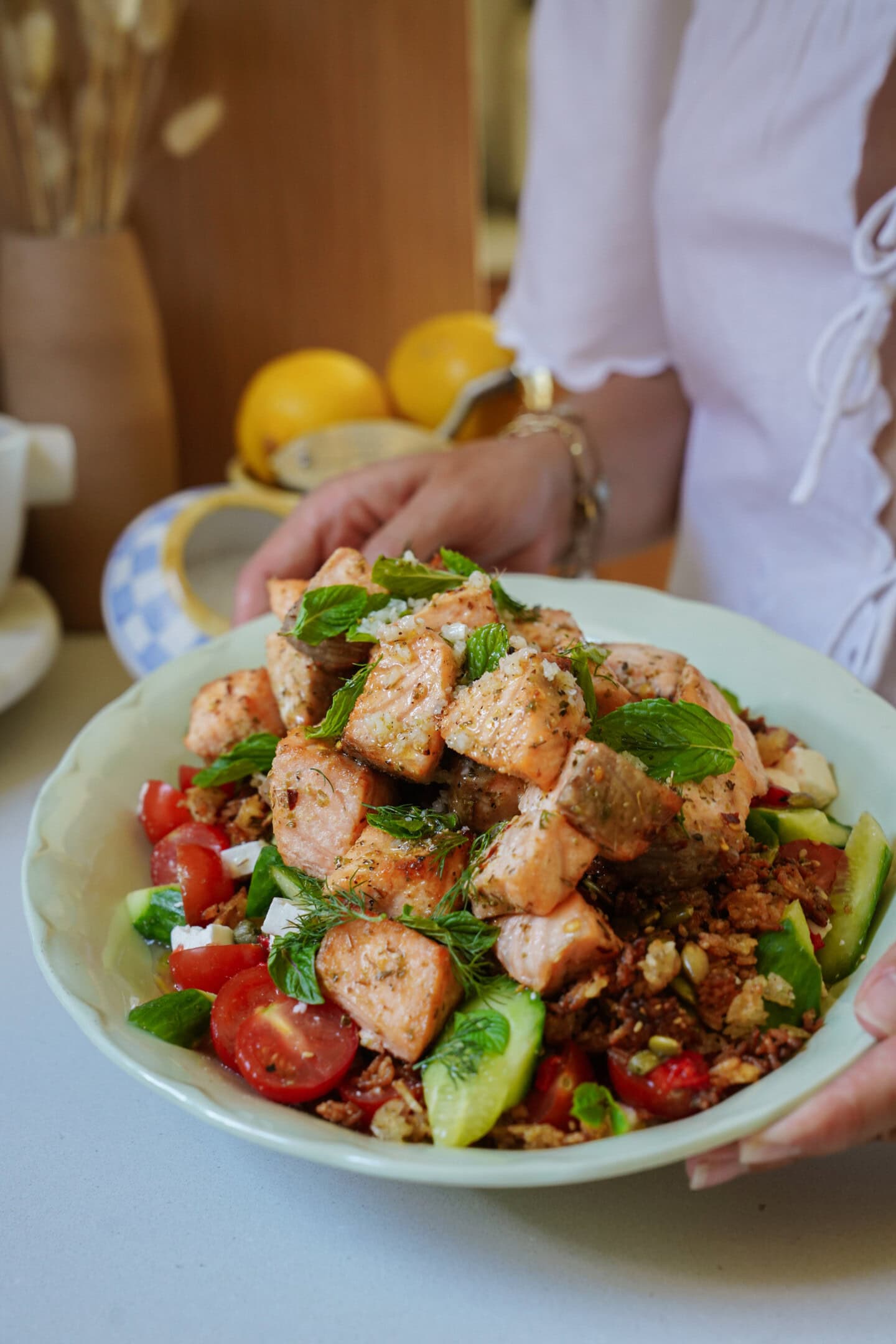 A person holding a bowl filled with a fresh salad topped with grilled salmon chunks, cherry tomatoes, cucumber slices, herbs, and feta cheese. Lemons and kitchen items are visible in the background.