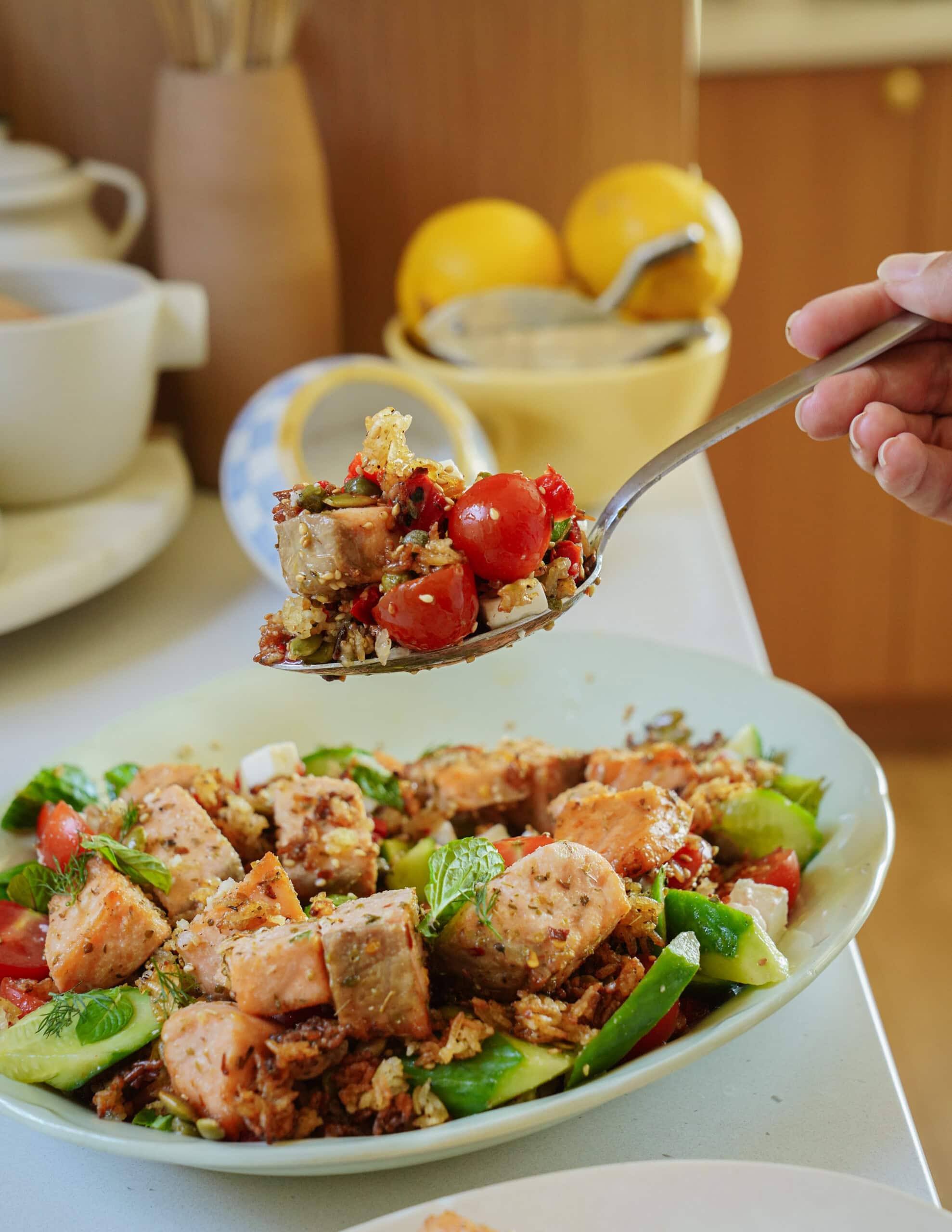 A close-up of a fresh salad with chunks of sausage, cherry tomatoes, cucumber, greens, and herbs on a plate, with a hand holding a spoonful of the salad above the dish. Lemons and kitchen items are in the background.