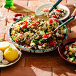 A large bowl of Mediterranean chickpea salad with tomatoes, cucumber, olives, and feta is being served with salad tongs. Lemons, garlic, and wine glasses are nearby on a terracotta-tiled table.