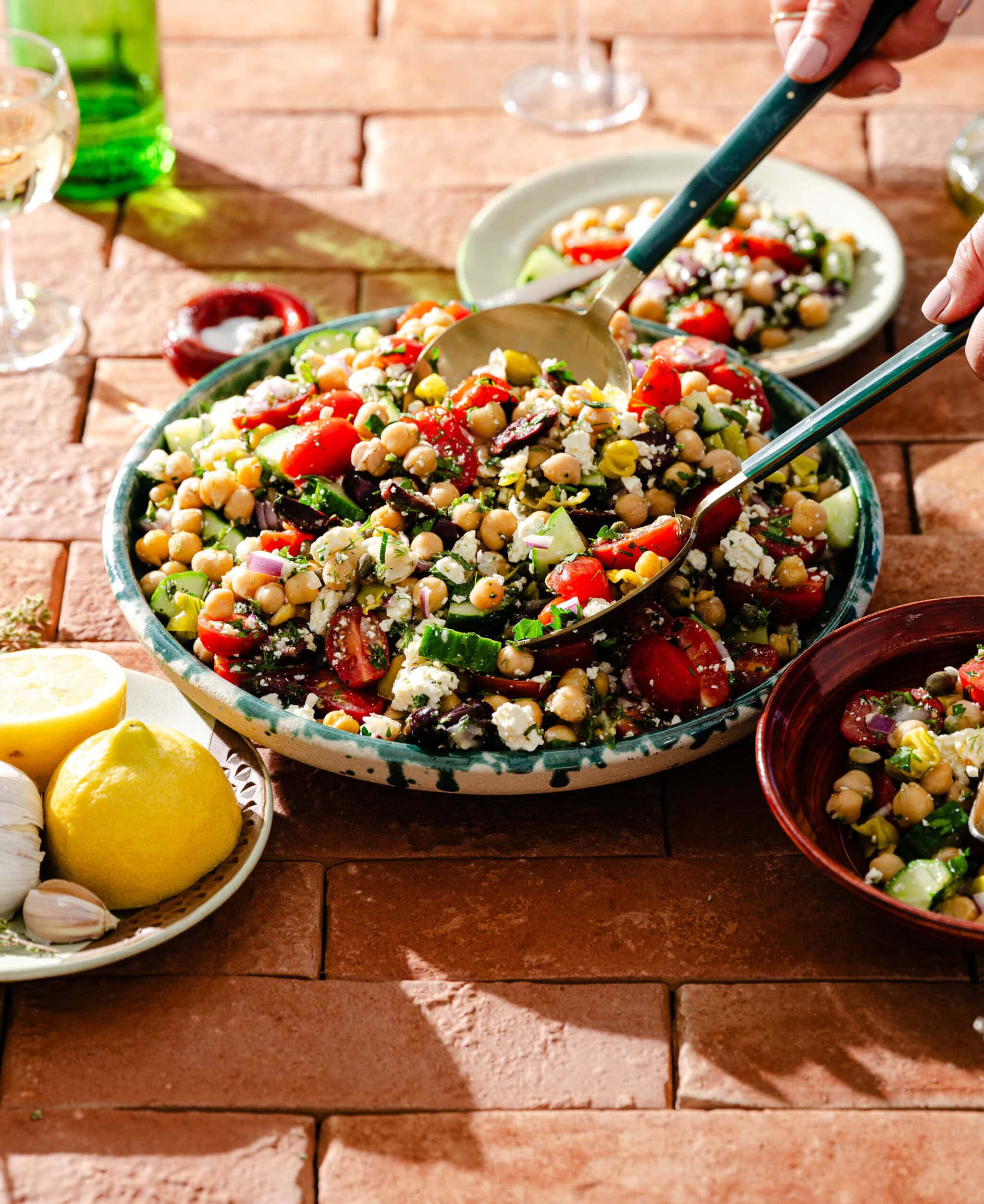 A large bowl of Mediterranean chickpea salad with tomatoes, cucumber, olives, and feta is being served with salad tongs. Lemons, garlic, and wine glasses are nearby on a terracotta-tiled table.