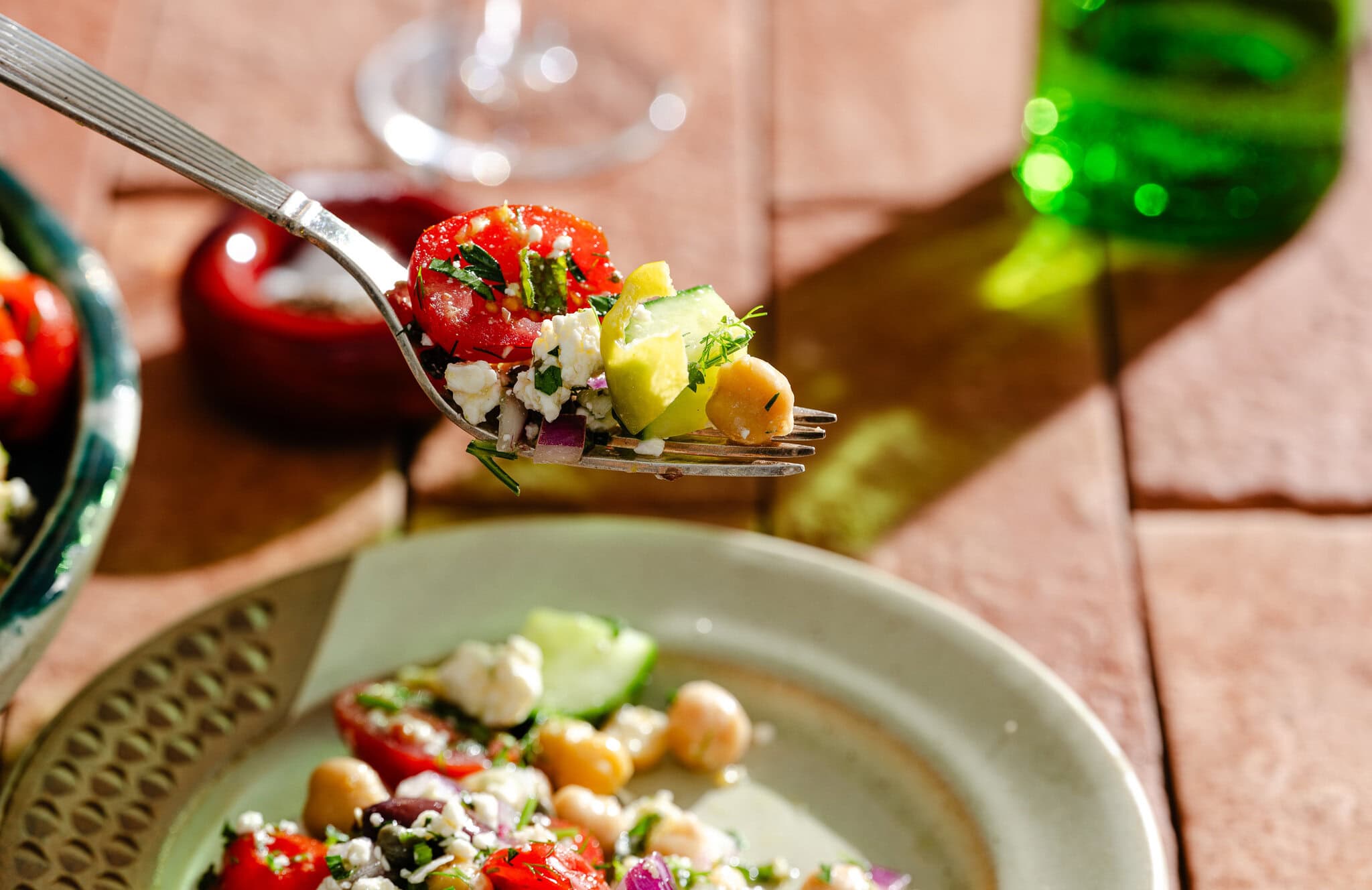 A close-up of a fork holding a bite of salad with tomato, cucumber, avocado, chickpeas, red onion, feta cheese, and herbs, above a plate with more salad on a sunlit table.