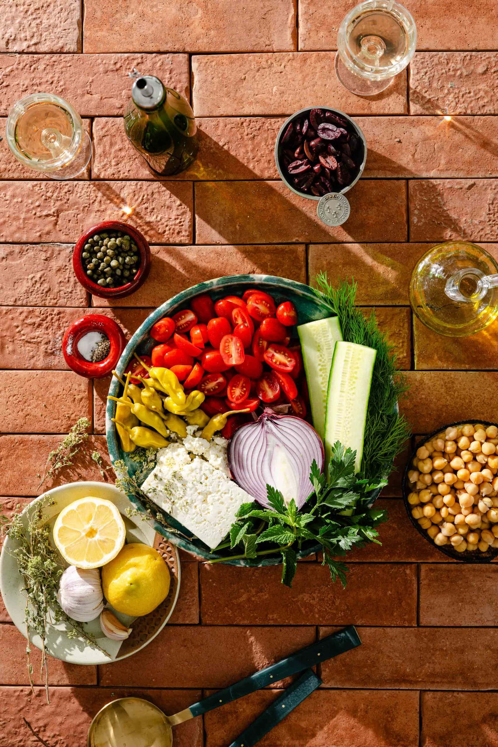 Overhead view of fresh Greek salad ingredients, including tomatoes, cucumber, red onion, feta, peppers, olives, chickpeas, capers, lemon, herbs, and garlic, arranged on a brick surface with glasses of white wine.