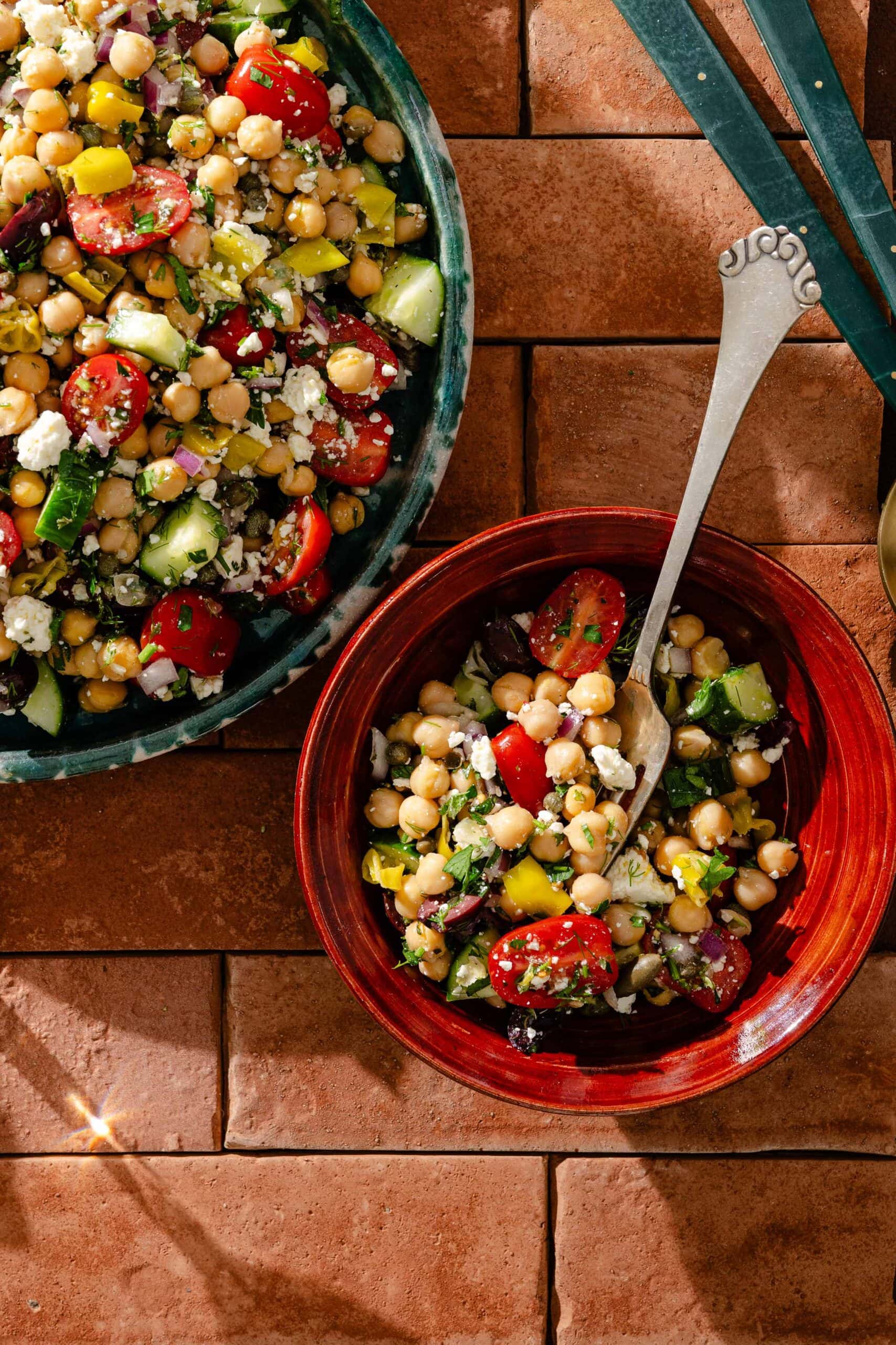 A colorful chickpea salad with cherry tomatoes, cucumbers, yellow bell peppers, red onions, feta cheese, and herbs served in a red bowl with a spoon, next to a larger serving bowl on a tiled surface.