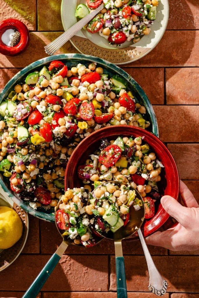 A colorful Mediterranean salad with chickpeas, cucumbers, cherry tomatoes, olives, red onions, and feta cheese is served in bowls on a tiled surface. A hand is scooping salad from a large bowl into a smaller red bowl.