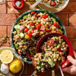 A large bowl of chickpea salad with cherry tomatoes, cucumber, red onion, feta cheese, herbs, and peppers. Nearby are a plate with sliced lemons and garlic, and a hand serving salad with a red spoon.