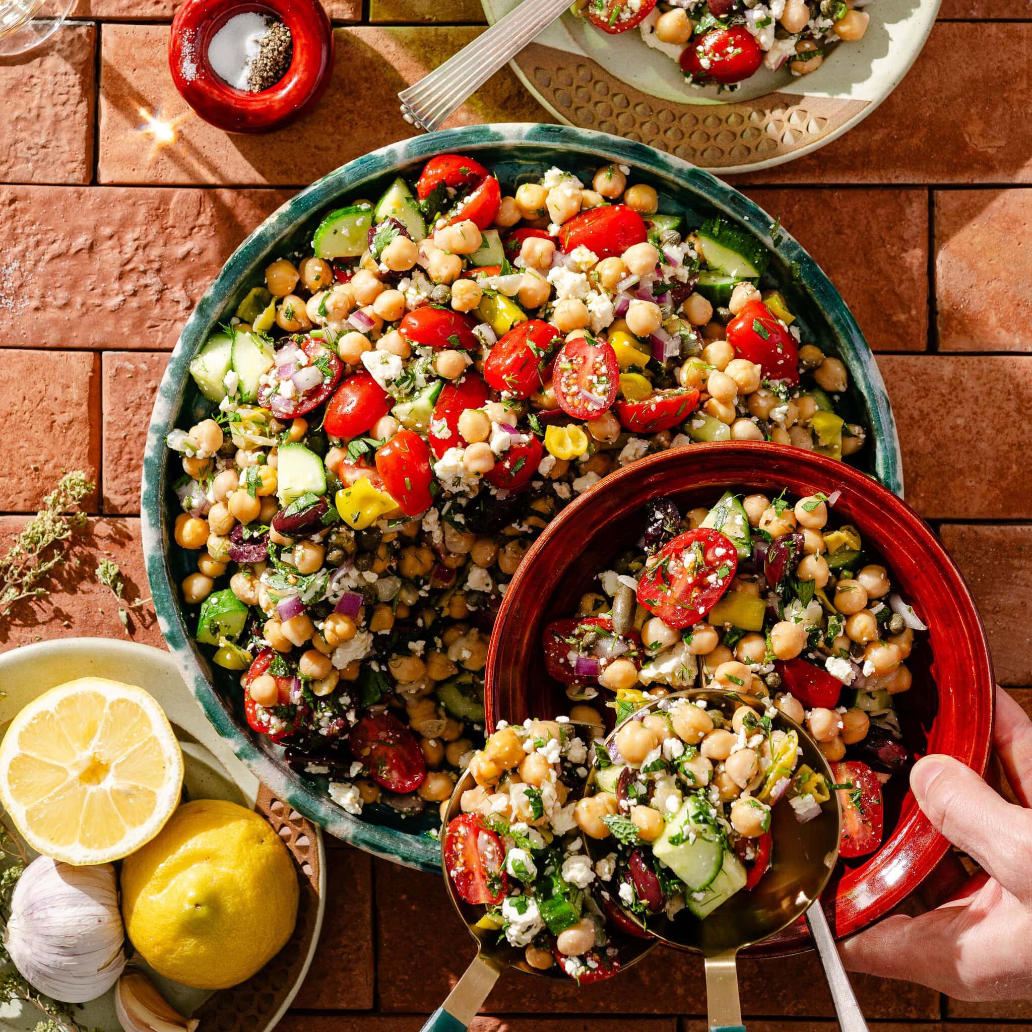 A large bowl of chickpea salad with cherry tomatoes, cucumber, red onion, feta cheese, herbs, and peppers. Nearby are a plate with sliced lemons and garlic, and a hand serving salad with a red spoon.