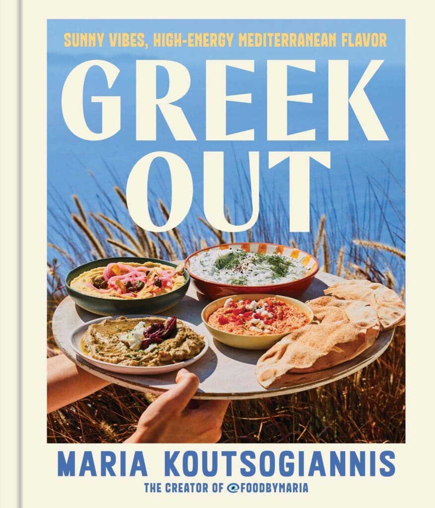 A person holds a tray with plates of Mediterranean food and pita bread outdoors, with the sea and tall grass in the background. The book title Greek Out and author Maria Koutsogiannis are displayed.