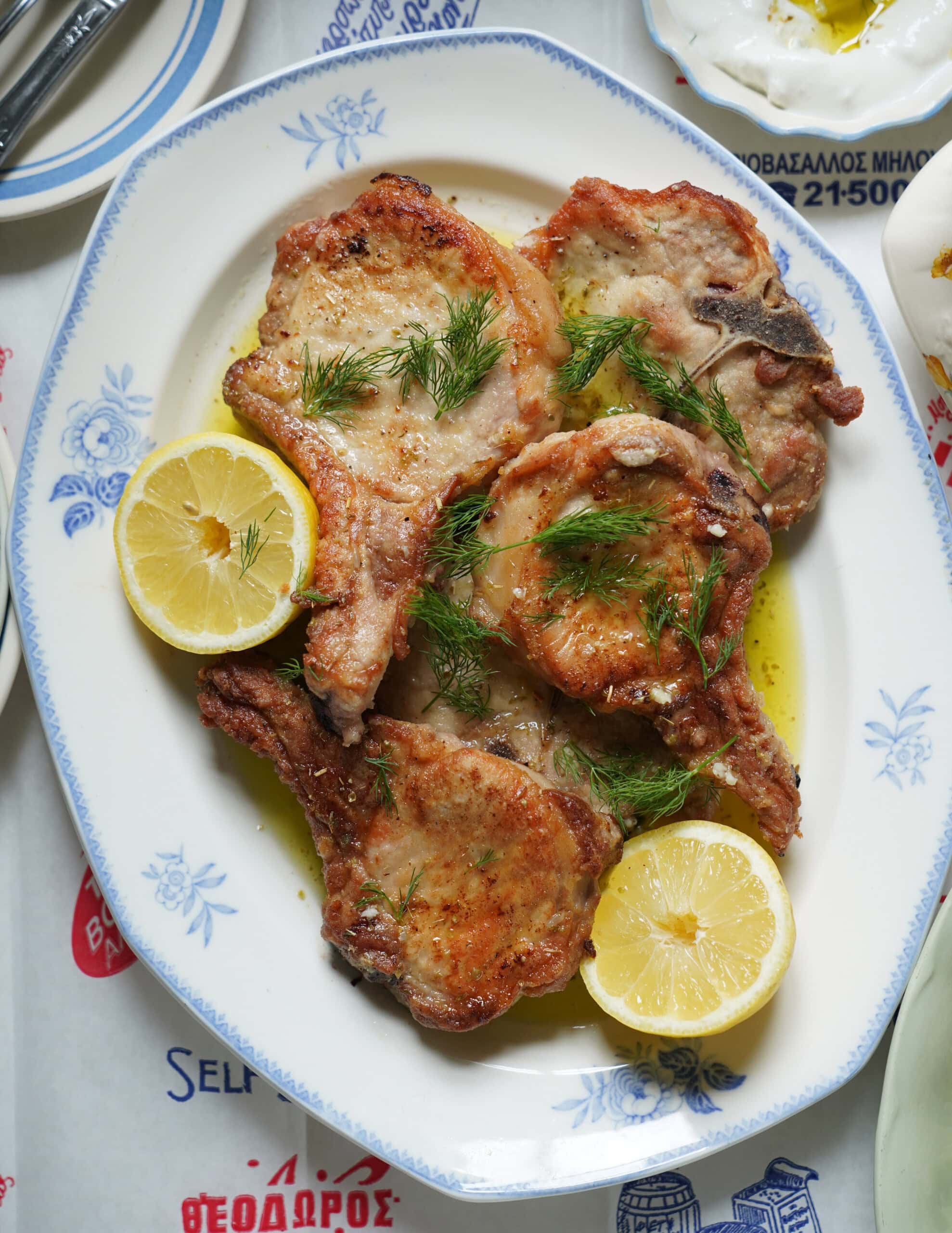 A plate of cooked pork chops garnished with fresh dill and served with lemon halves, presented on a white dish with a blue floral border.