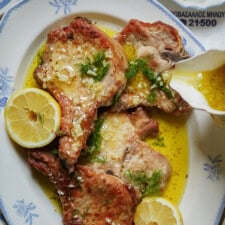 Grilled pork chops garnished with fresh dill and lemon slices on a white plate, with a rich yellow sauce being poured over them from a small pitcher.