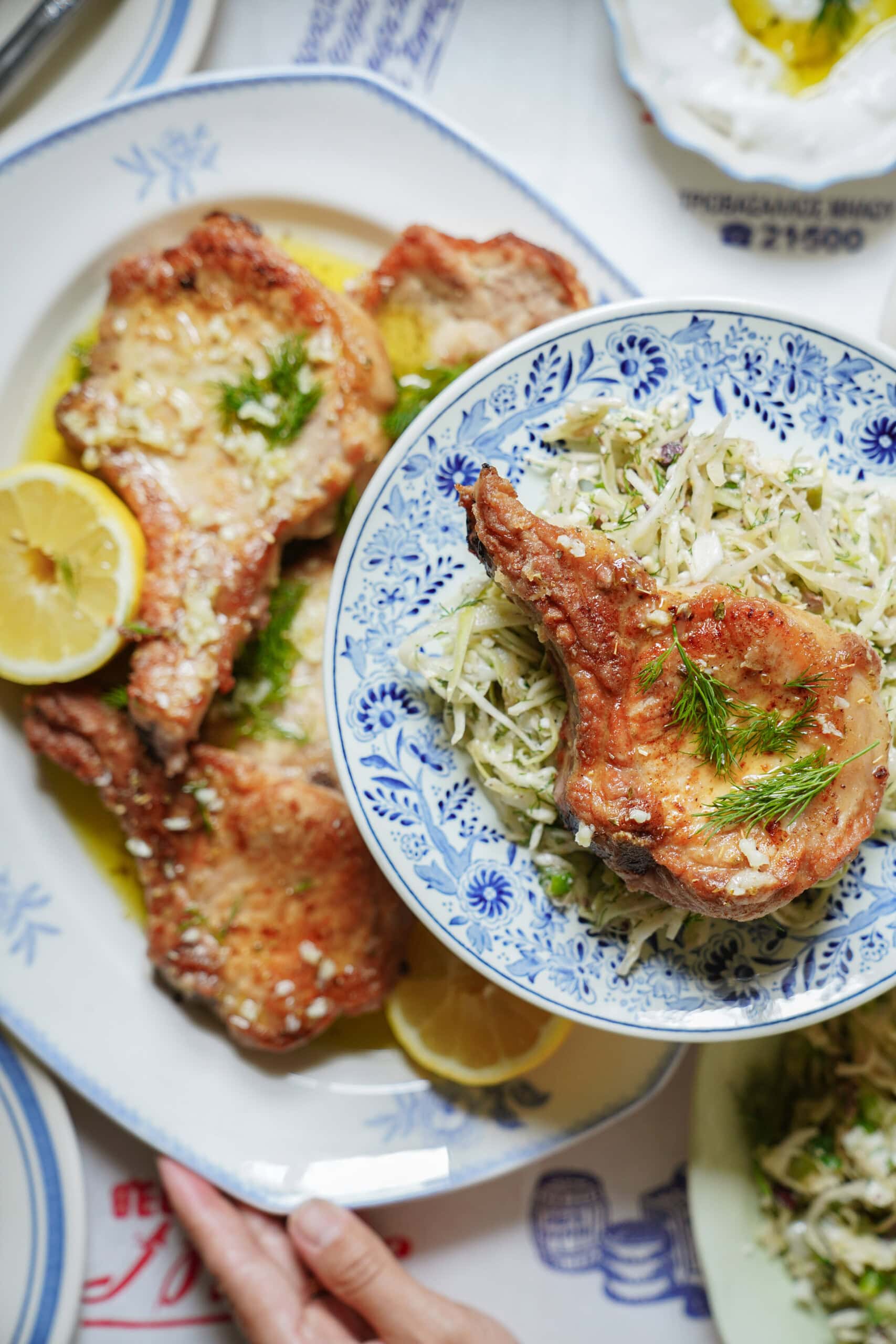 A plate of golden-brown pork chops garnished with dill and lemon slices is served with shredded cabbage salad on a blue and white bowl, all on a patterned tablecloth.